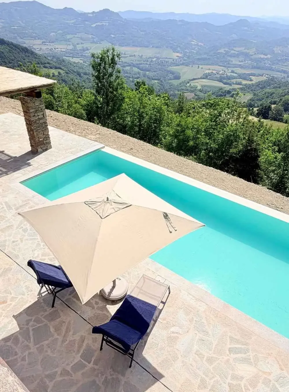 A swimming pool on a patio with lounge chairs, a large umbrella, and a scenic view of green hills and valleys in the distance.