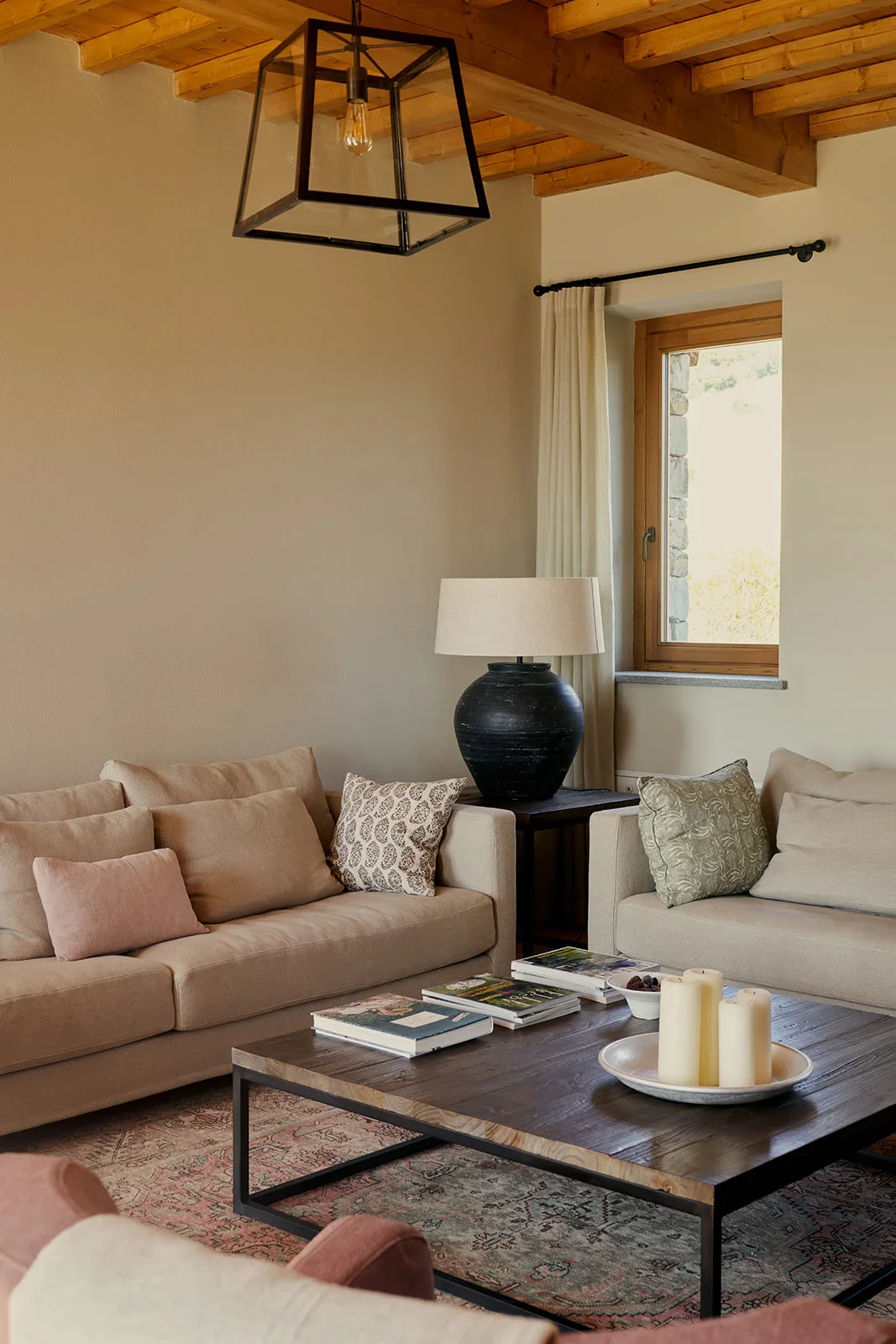 Living room with beige sofas, patterned cushions, a wooden coffee table with candles and magazines, a black table lamp on a side table, a window with a wooden frame and cream curtains, and a modern geometric pendant light hanging from a wooden ceilin