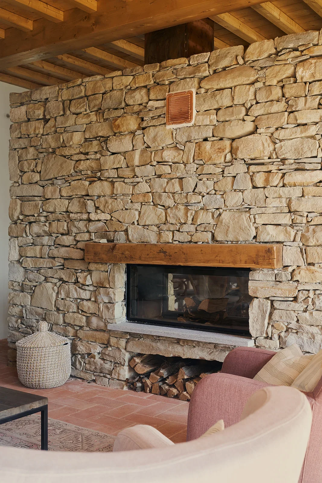A cozy living room corner featuring a stone fireplace with a wooden mantle, a basket on the floor, and a pink armchair with a beige pillow.