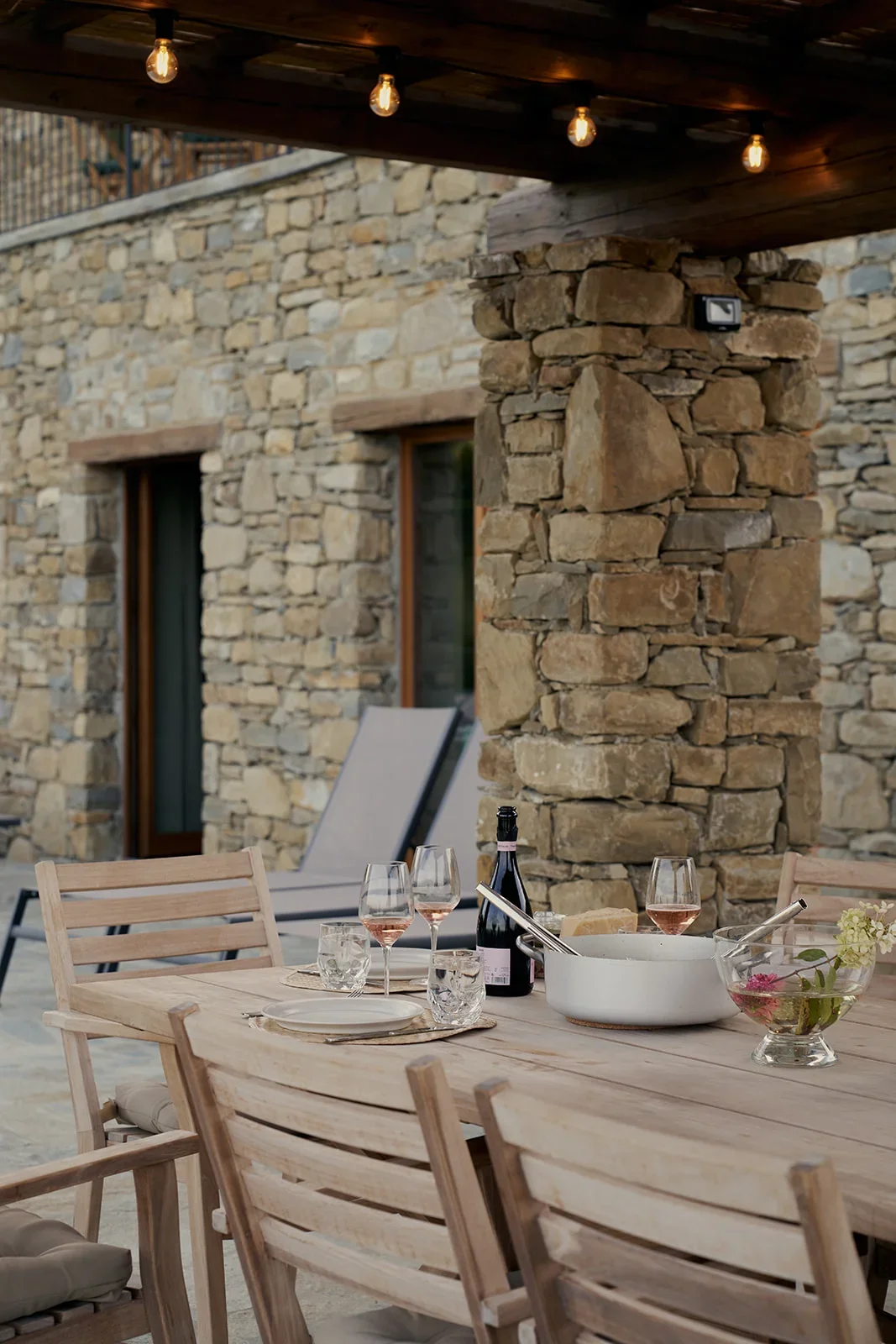 An outdoor dining area with a wooden table set with wine glasses, a wine bottle, and a bowl of flowers, set against a rustic stone wall and wooden beams with string lights overhead.