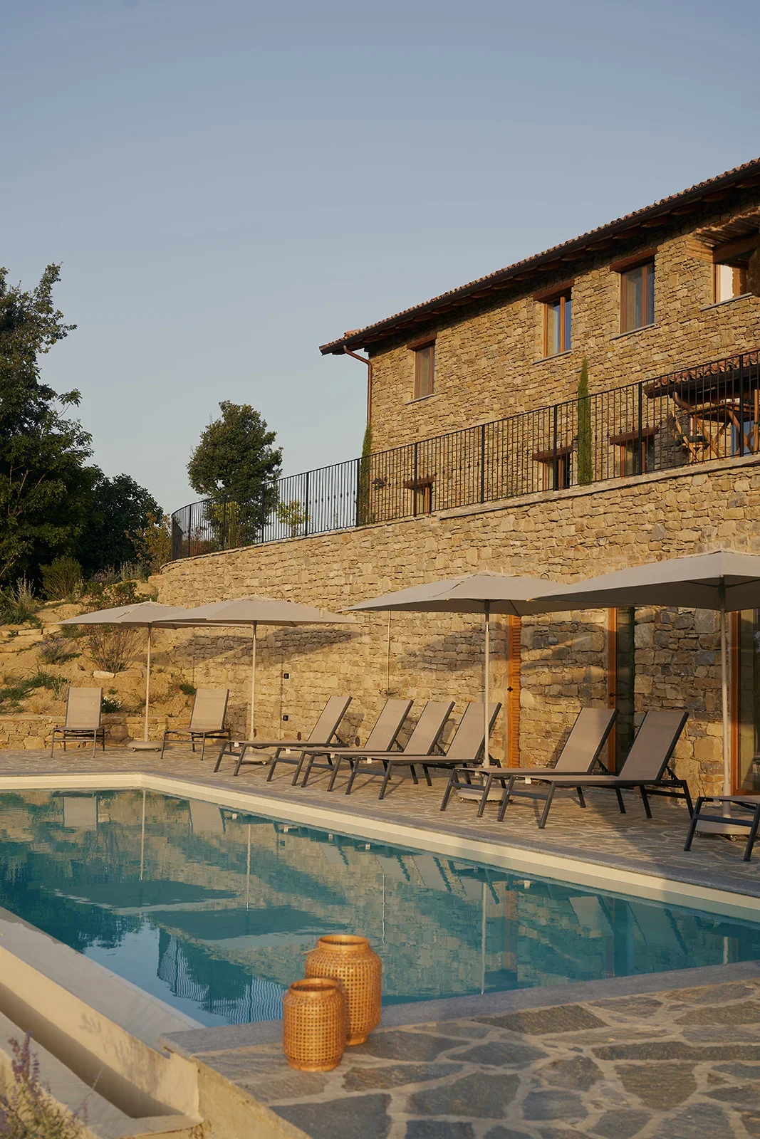 Luxury house with stone exterior and large windows, outdoor swimming pool with lounge chairs and umbrellas, and decorative vases, set against a clear sky.