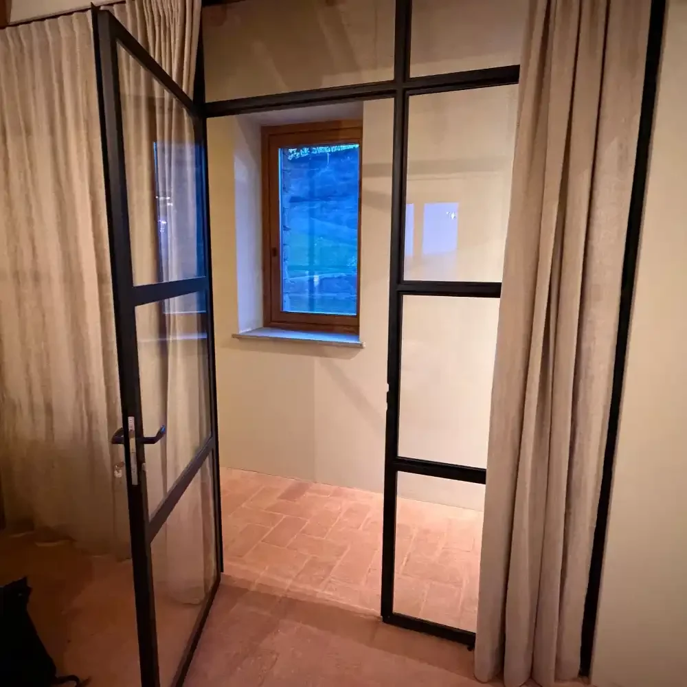 Interior view of a small room with a window, metal-framed glass door, beige curtains, and terracotta tile flooring.