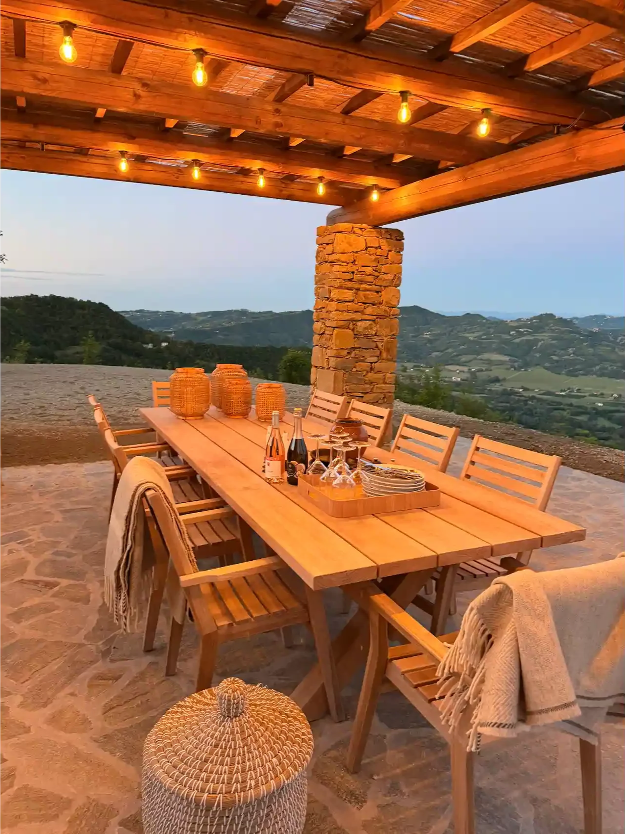An outdoor dining area with a long wooden table and chairs, decorated with lanterns, wine bottles, glasses, and plates. The setting overlooks a scenic landscape with rolling hills and a clear sky at dusk.