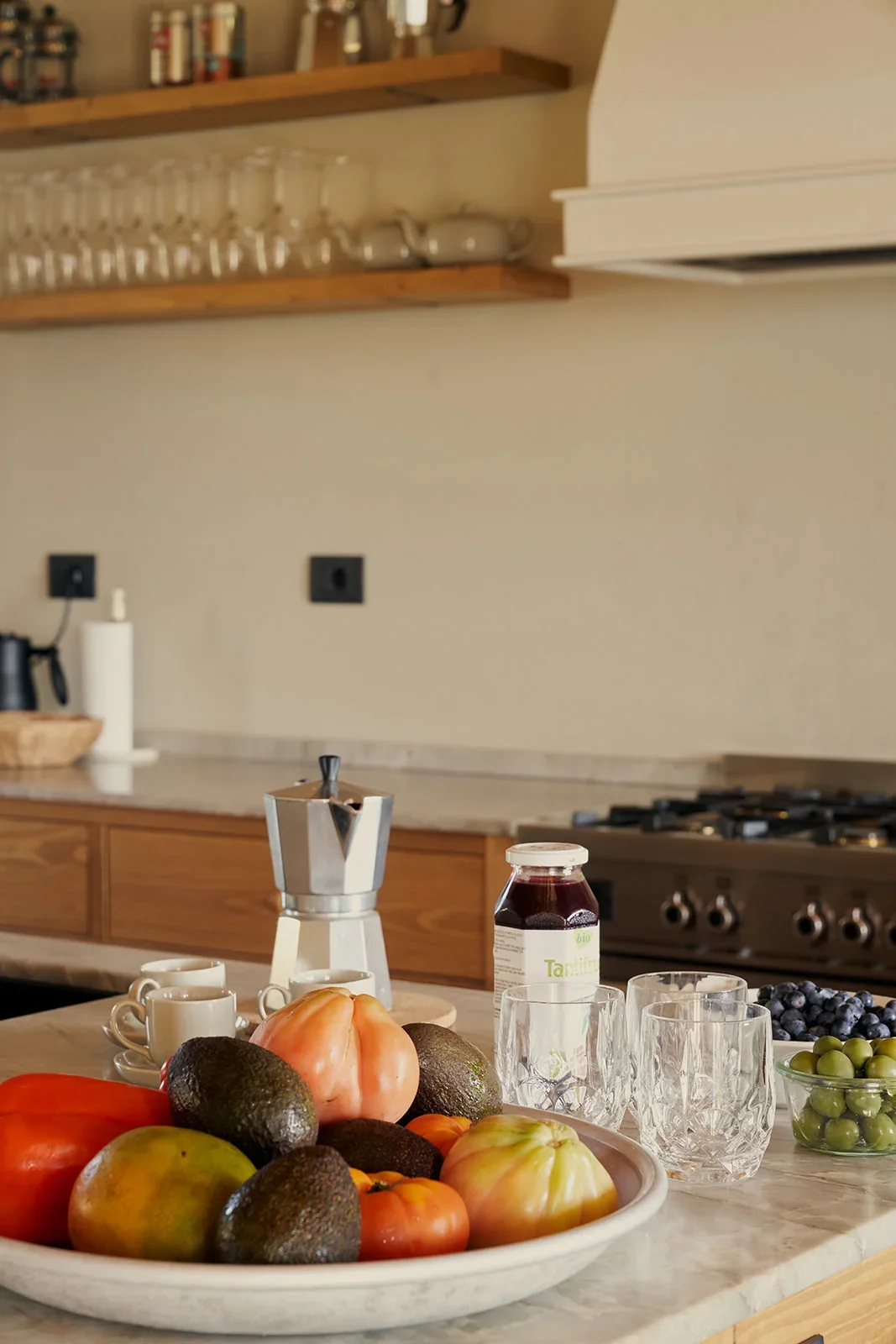 Kitchen counter with a bowl of tomatoes, avocados, and other fruits, a jar of jam, four empty glasses, a moka pot, three cups, and a small bowl of grapes.
