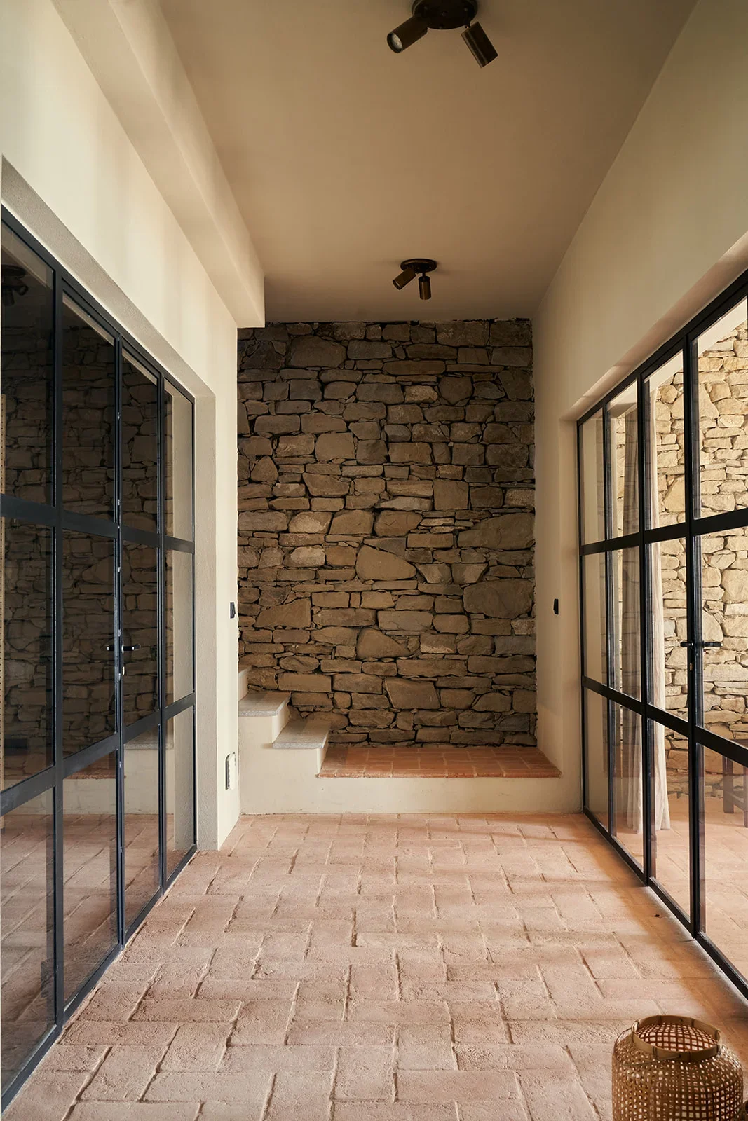 Room with brick tile flooring, stone wall at the end, white walls, black metal framed glass sliding doors on both sides, and a small lantern on the floor.