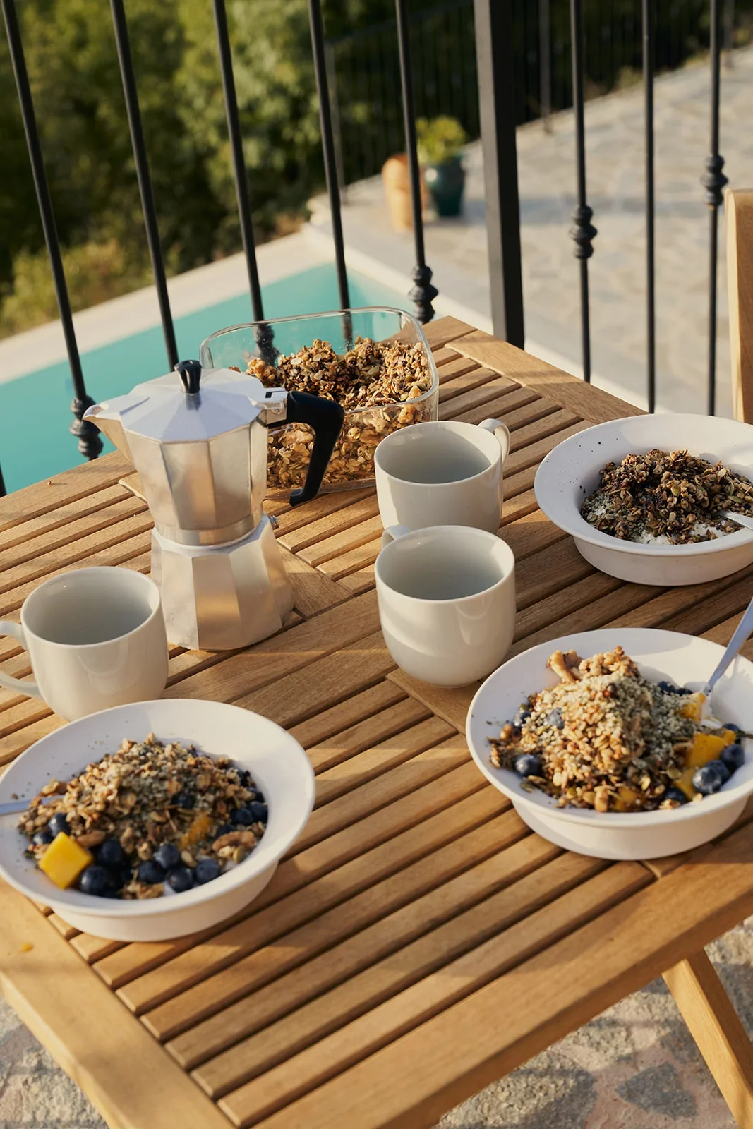 A wooden outdoor table set with bowls of granola and blueberries, cups of coffee, a stovetop espresso maker, and a glass jar of granola, with a pool and greenery in the background.