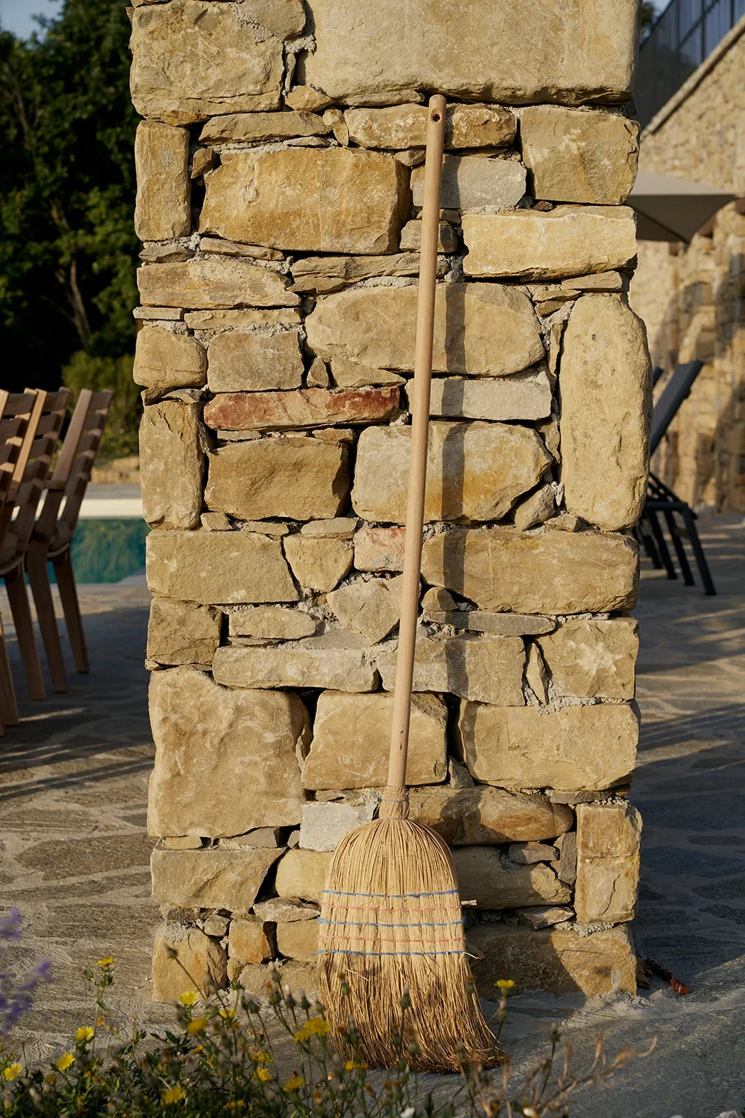 A broom leaning against a stone pillar outdoors, with a patio and outdoor furniture visible in the background.