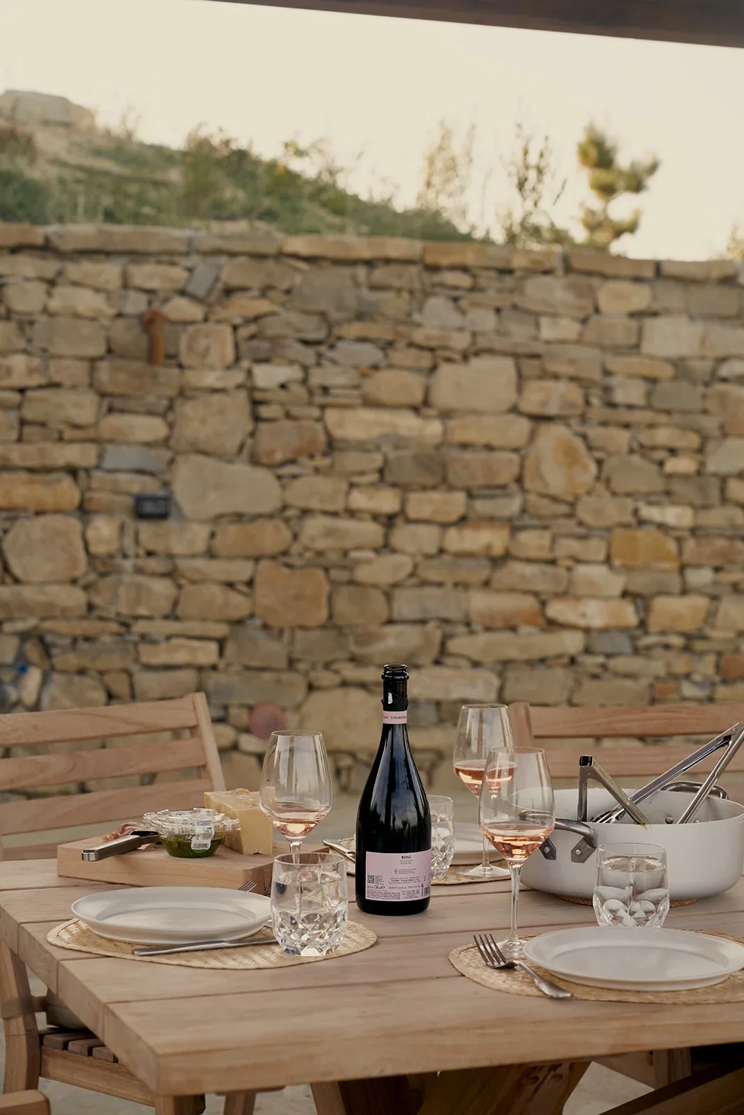A wooden outdoor dining table set for four with wine glasses, a bottle of rosé wine, a cheese platter, and cutlery, with a stone wall and trees in the background.
