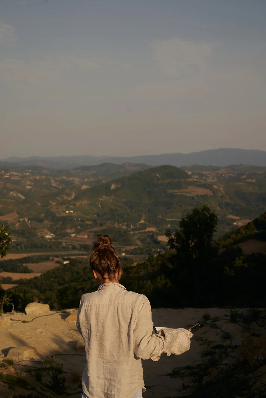 A woman with brown hair in a bun, wearing a light-colored jacket, standing outdoors at sunset, overlooking a landscape of rolling hills and valleys.