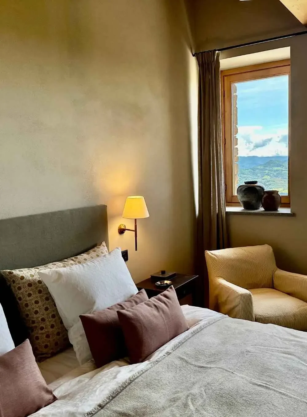 A bedroom with a bed, pillows, a wall-mounted lamp, a bedside table with a tray, a beige armchair, and a window with a view of mountains and clouds, decorated with large ceramic vases.