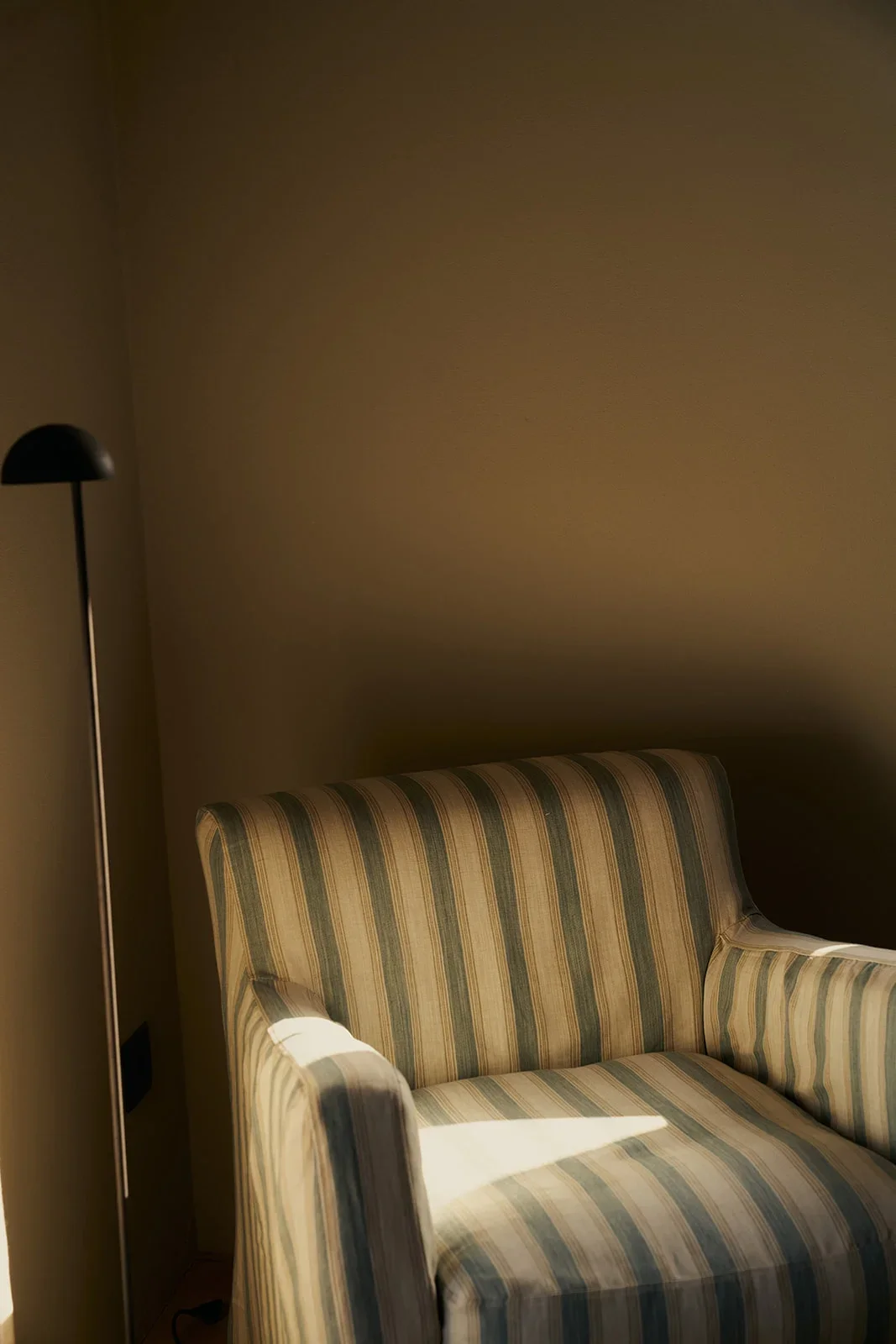 A room corner with a striped sofa, beige walls, and a black floor lamp with a round shade, partially illuminated by sunlight.