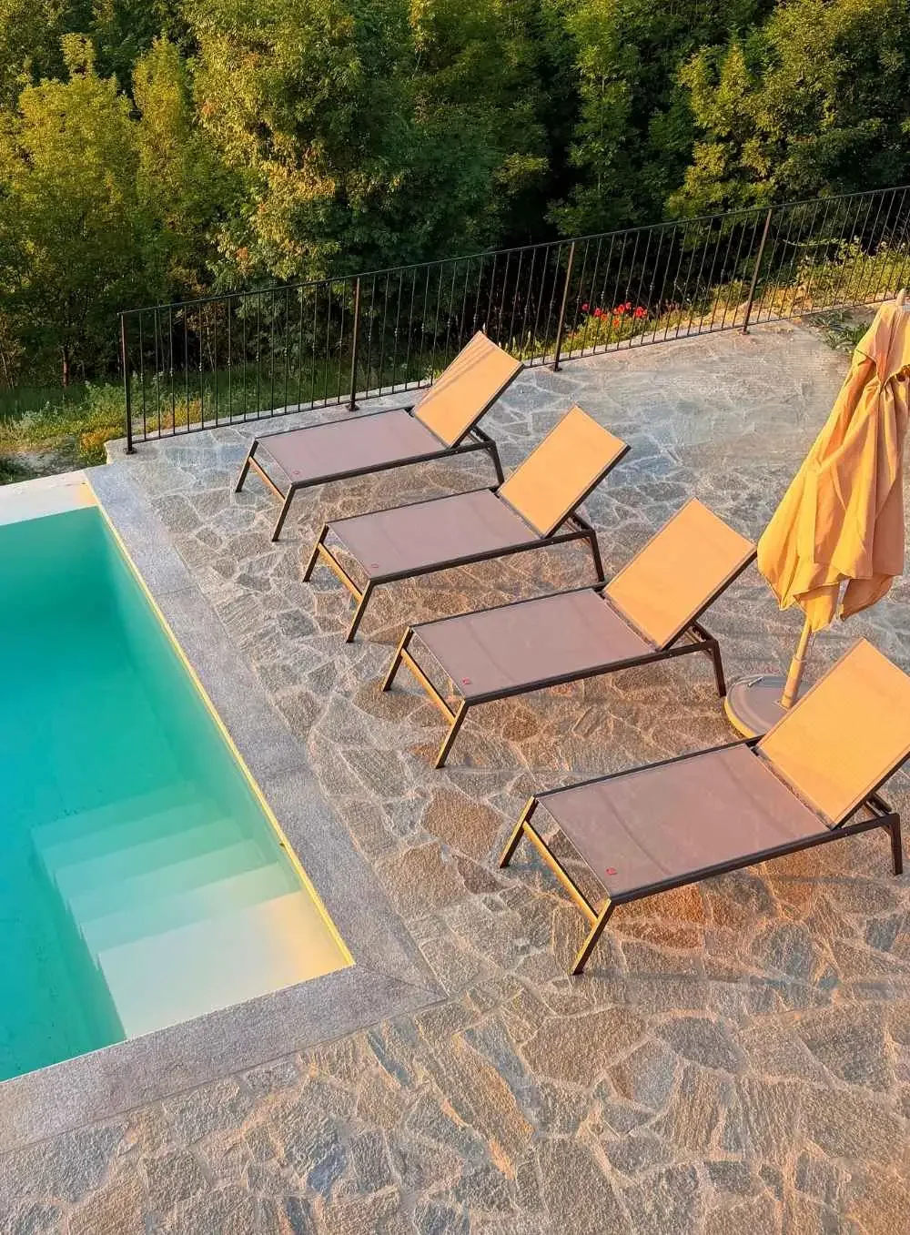 Four lounge chairs placed on a stone-tiled poolside patio with an umbrella, overlooking trees and greenery.