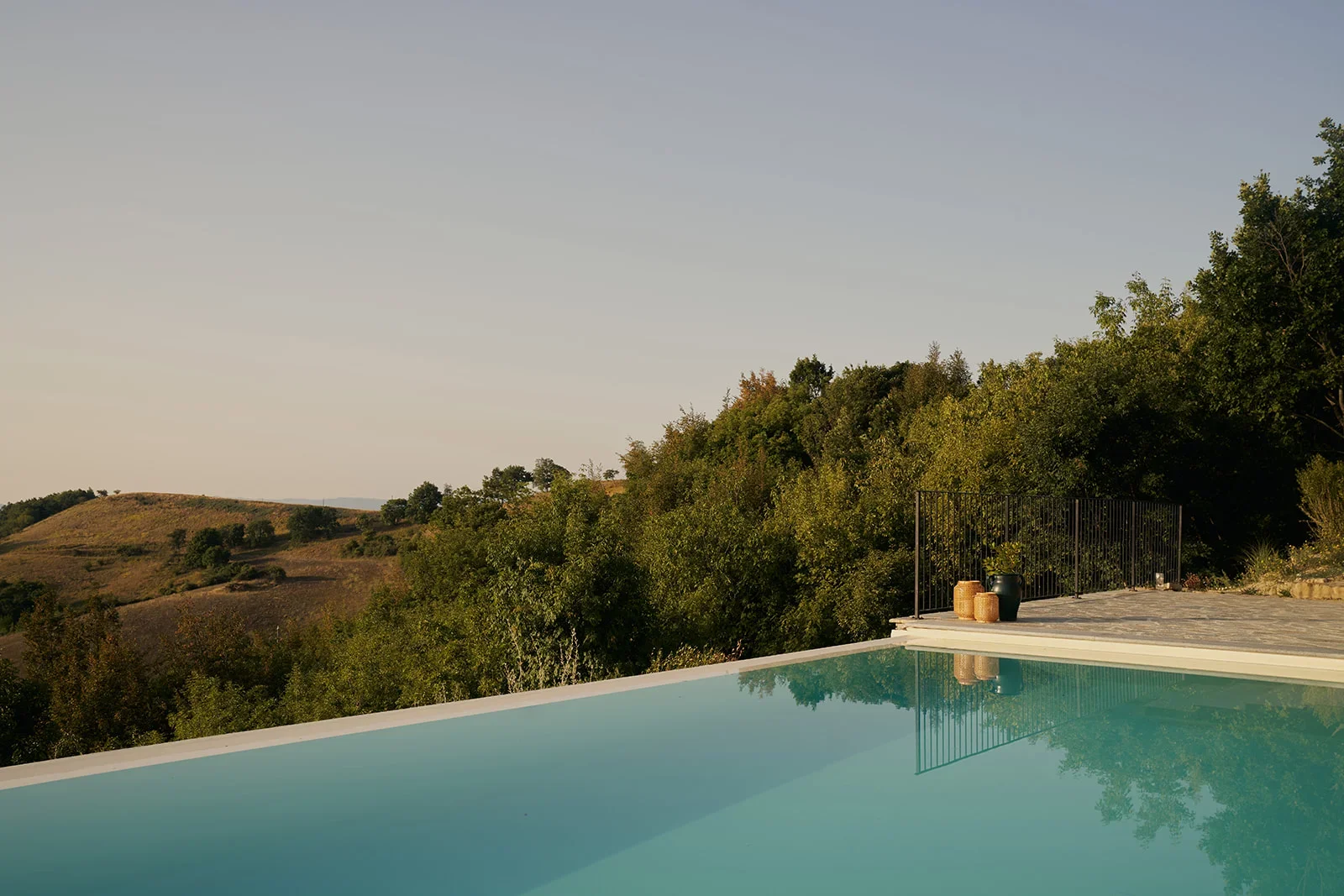 An outdoor pool with a scenic view of rolling hills and trees, featuring a deck with potted plants and decorative baskets at sunset.