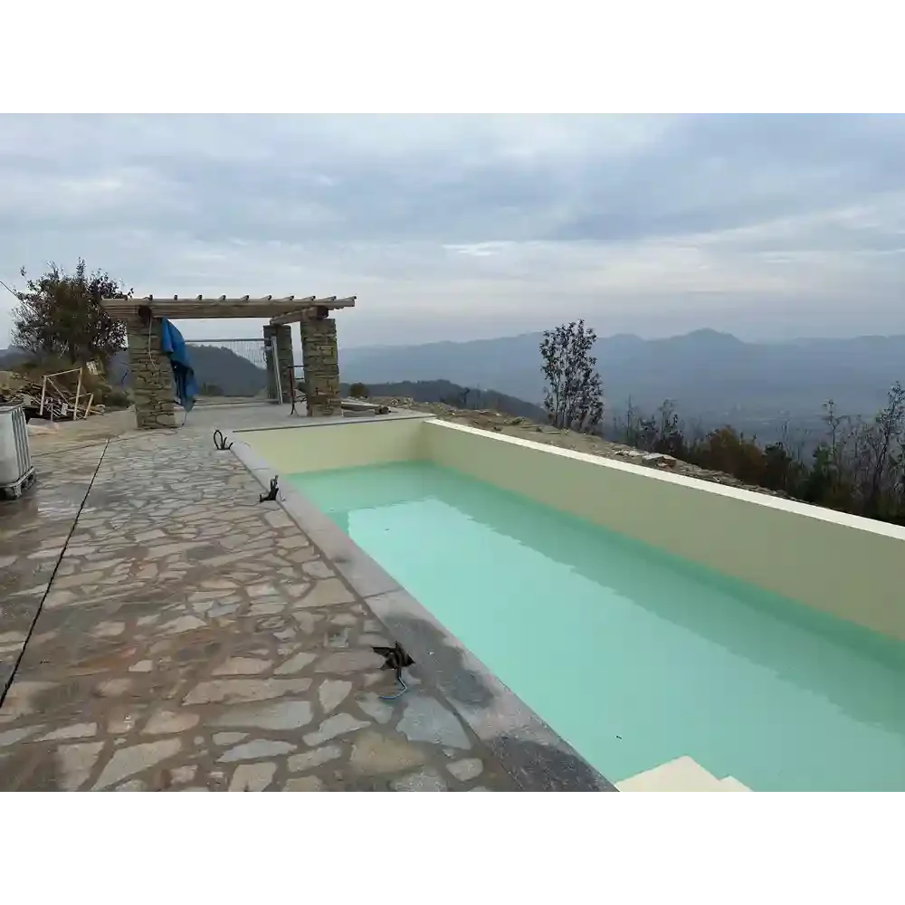 An empty swimming pool on a mountain terrace with a stone floor, overlooking a foggy mountain landscape under a cloudy sky.