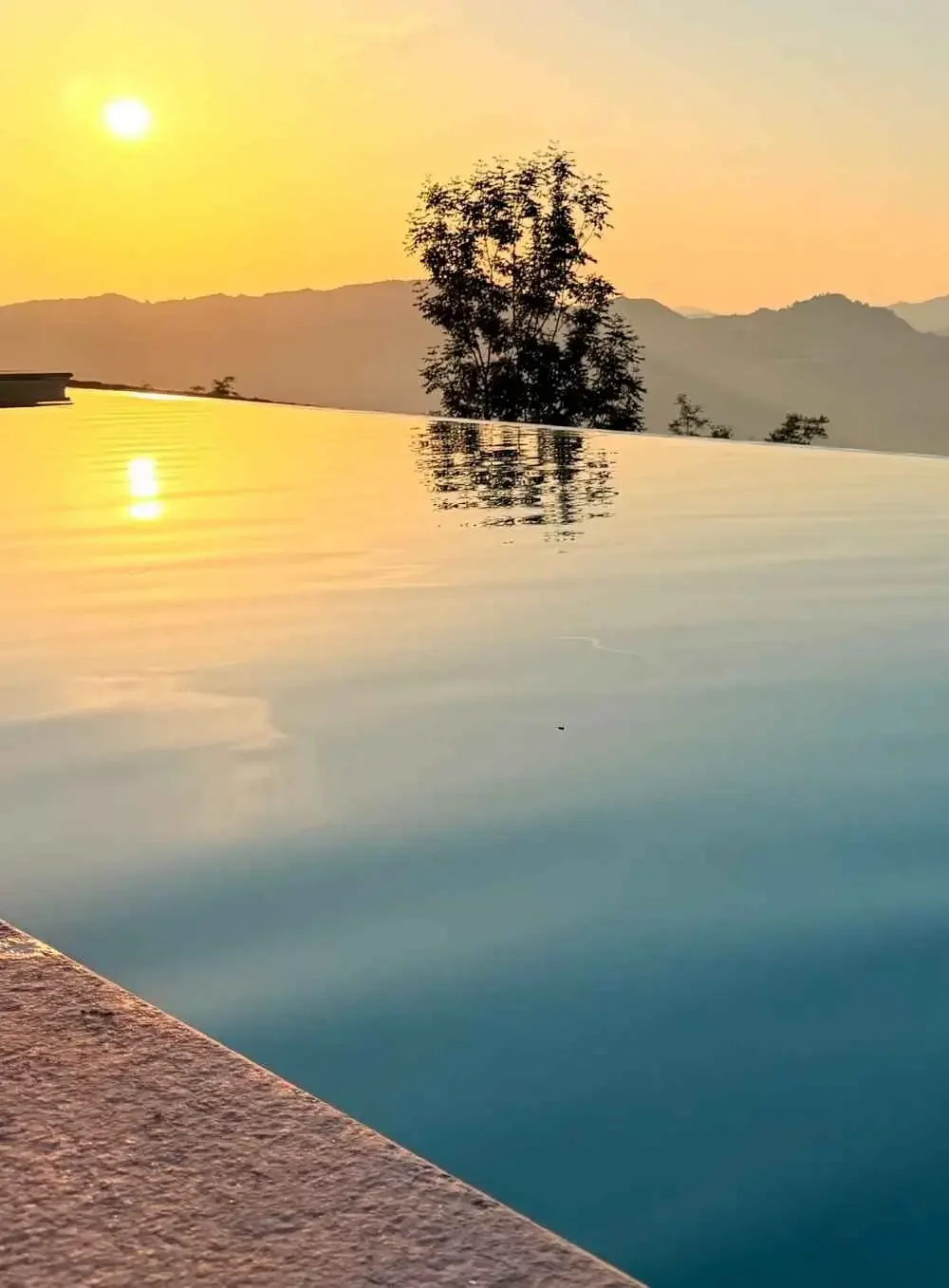 Sunset over a mountain range with a tree in silhouette reflected in a pool of water, with the pool's edge in the foreground.