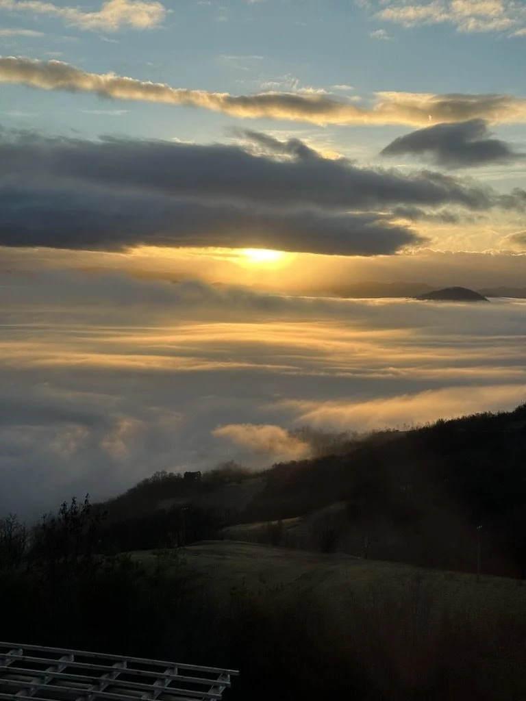 Sunset over clouds with a mountainous landscape below, some trees and hills, and a bright sky with scattered clouds.