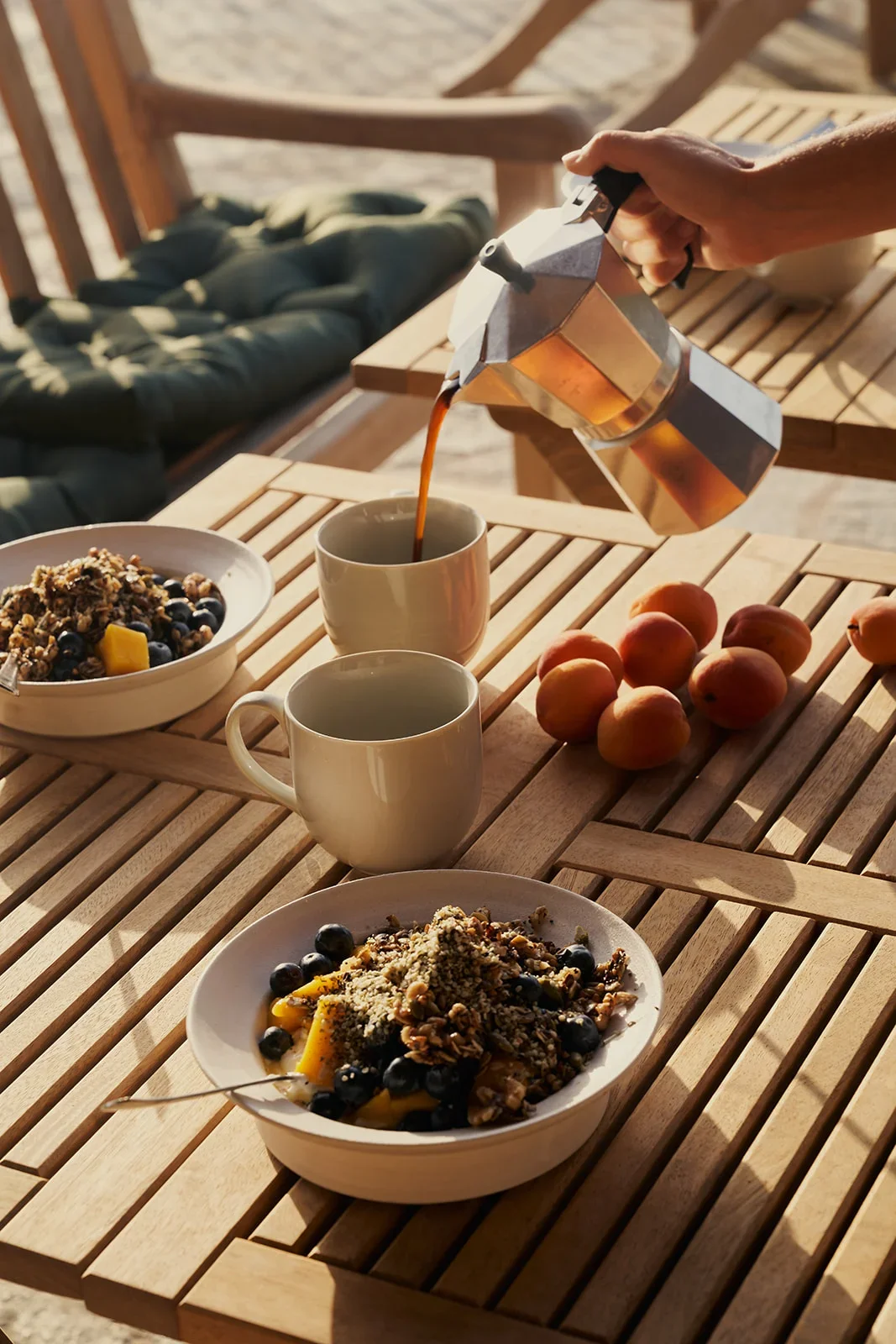 Pouring coffee into a cup on a wooden outdoor table with bowls of granola, blueberries, peaches, and a bowl of mixed fruit and granola in the foreground, with a beach chair and sand in the background.