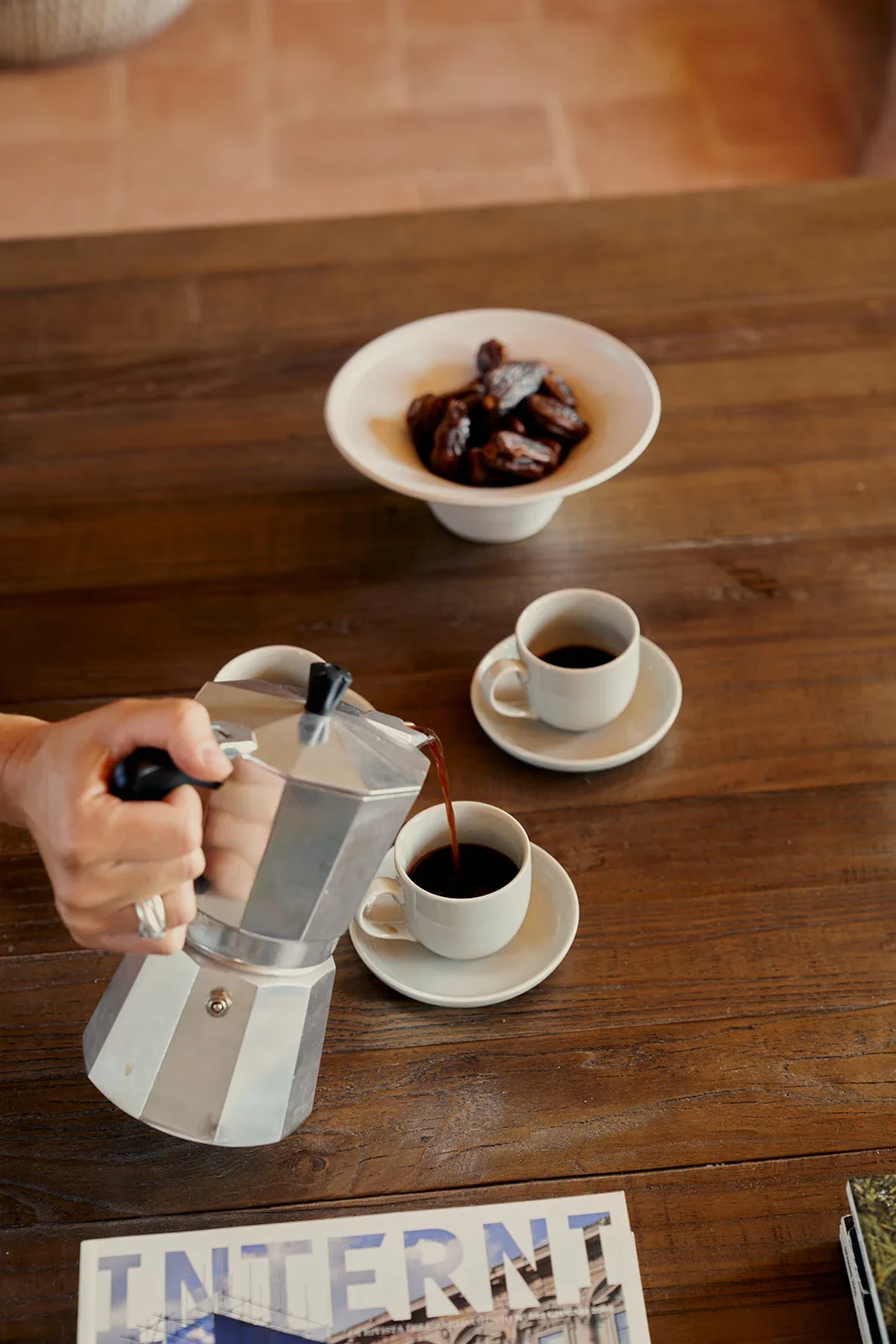 A person pouring coffee from a moka pot into two cups, with a bowl of dates and a partially visible magazine on a wooden table.