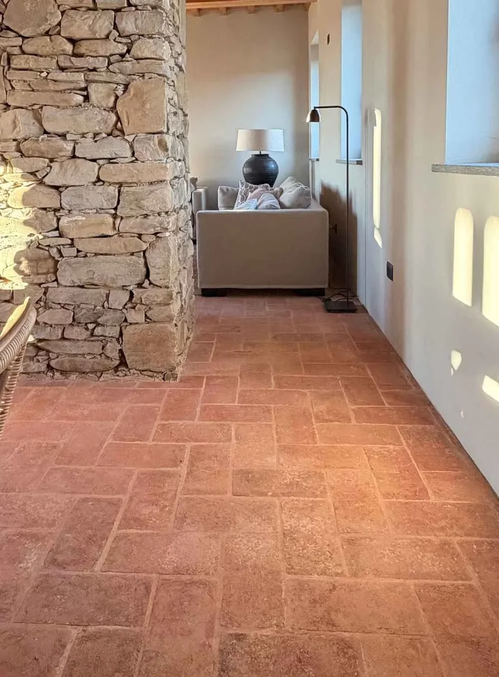 Interior of a house with a stone wall on the left, terracotta tile floor, white walls, and a cozy sitting area with cushions, a table lamp, and a floor lamp in the background.