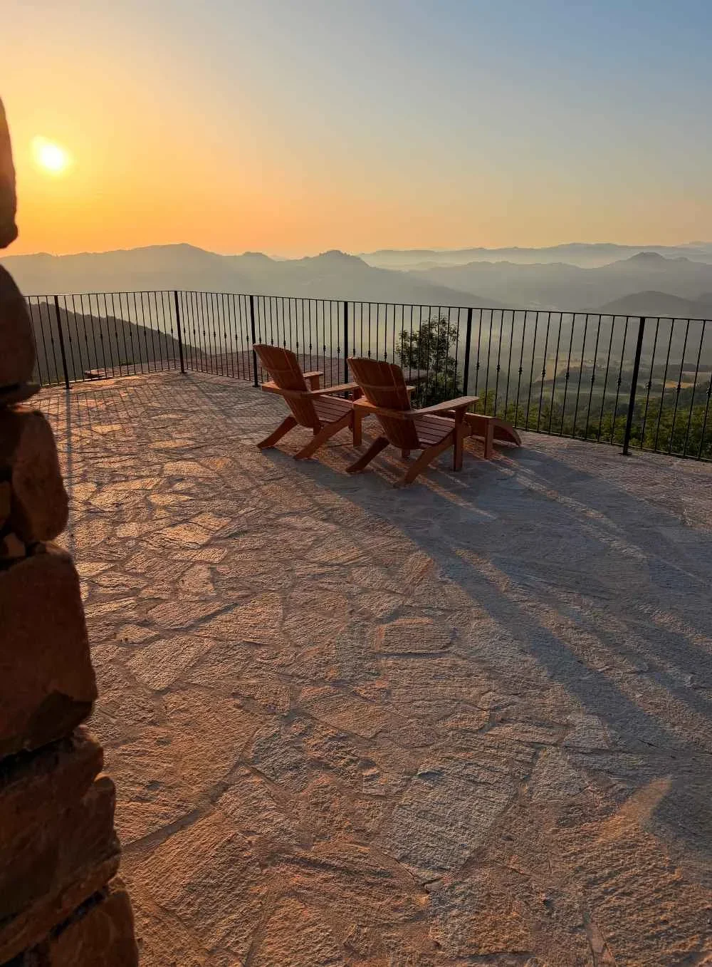 Sunset view from a stone balcony with two wooden lounge chairs overlooking a layered mountain landscape in the distance.