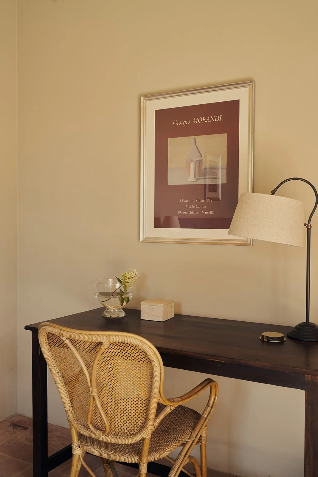 A cozy corner with a wooden desk, wicker chair, framed art on the wall, a table lamp, a small flower arrangement in a glass bowl, and a box on the desk.