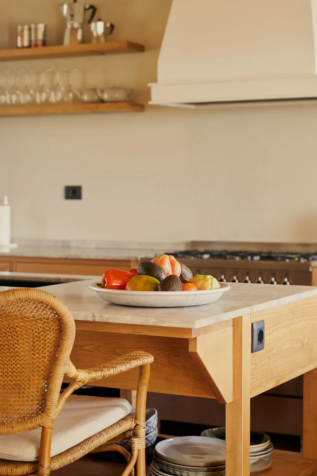 A kitchen with wooden furniture, a marble countertop, and a white bowl of assorted fruits including avocados, peppers, and tomatoes. There is a rattan chair in the foreground and shelves with glassware and books in the background.