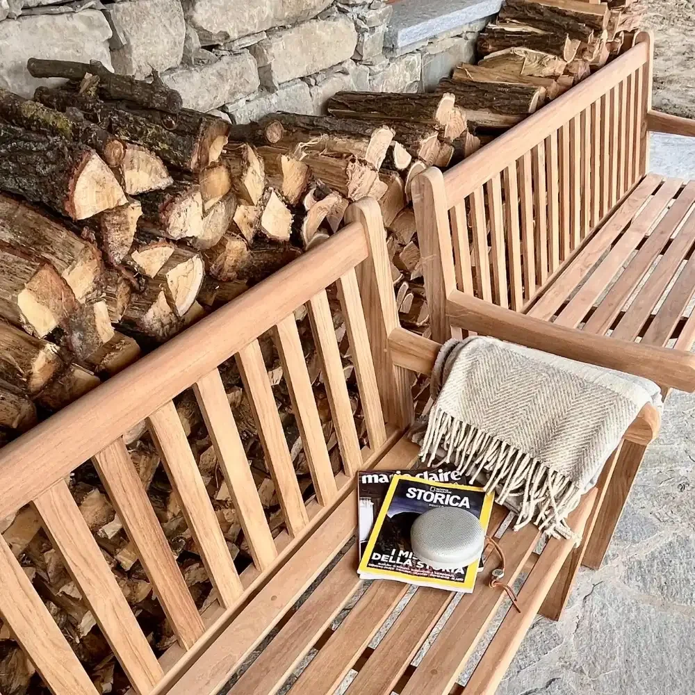 Wooden benches with a stack of magazines, a small gray speaker, and a woven blanket, against a stone firewood storage wall.