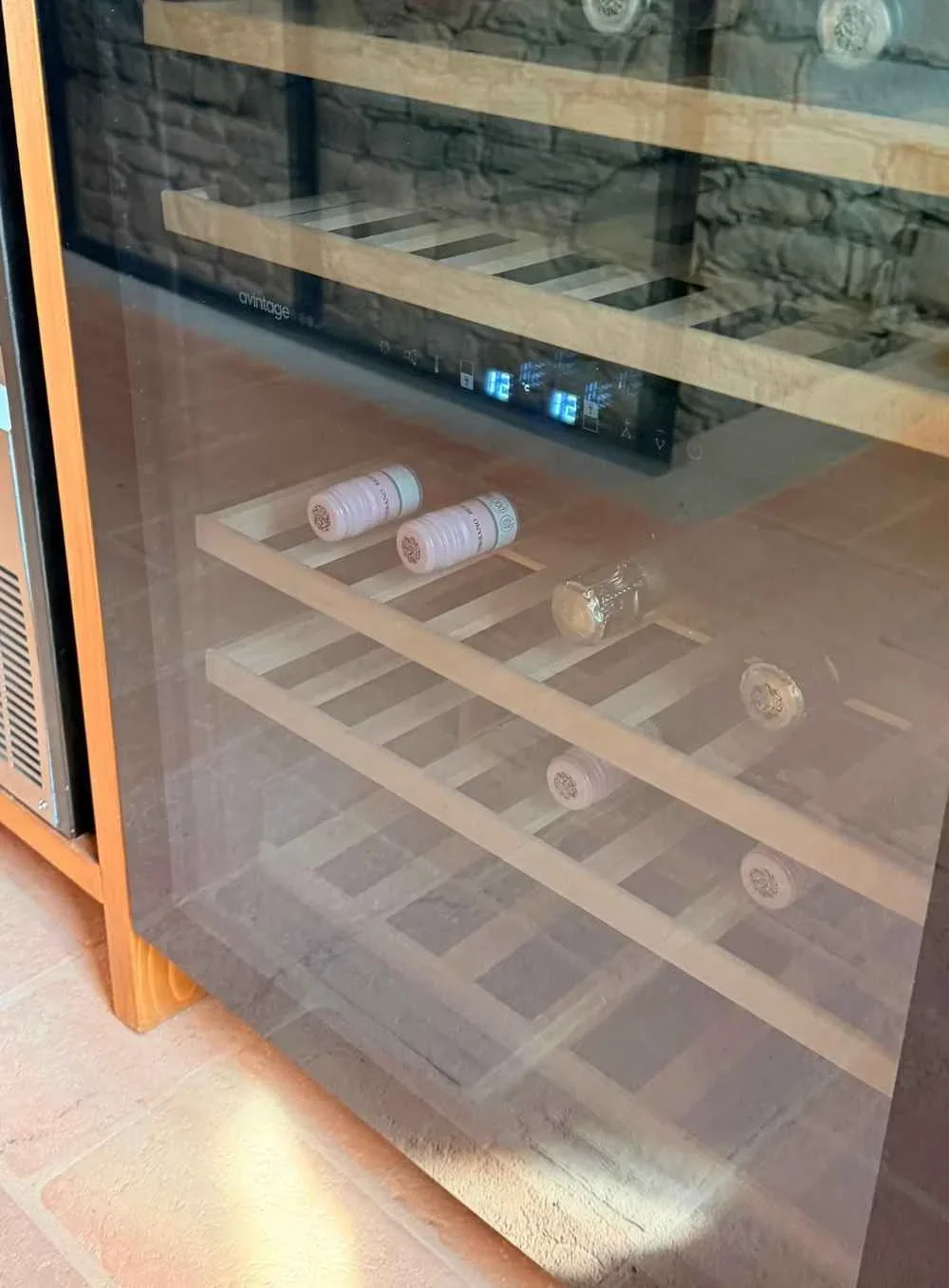 Empty wine cooler with a digital temperature display showing 12 degrees Celsius, containing a few wine bottles lying on their sides, with some beverage cans on the lower shelf behind the glass door.