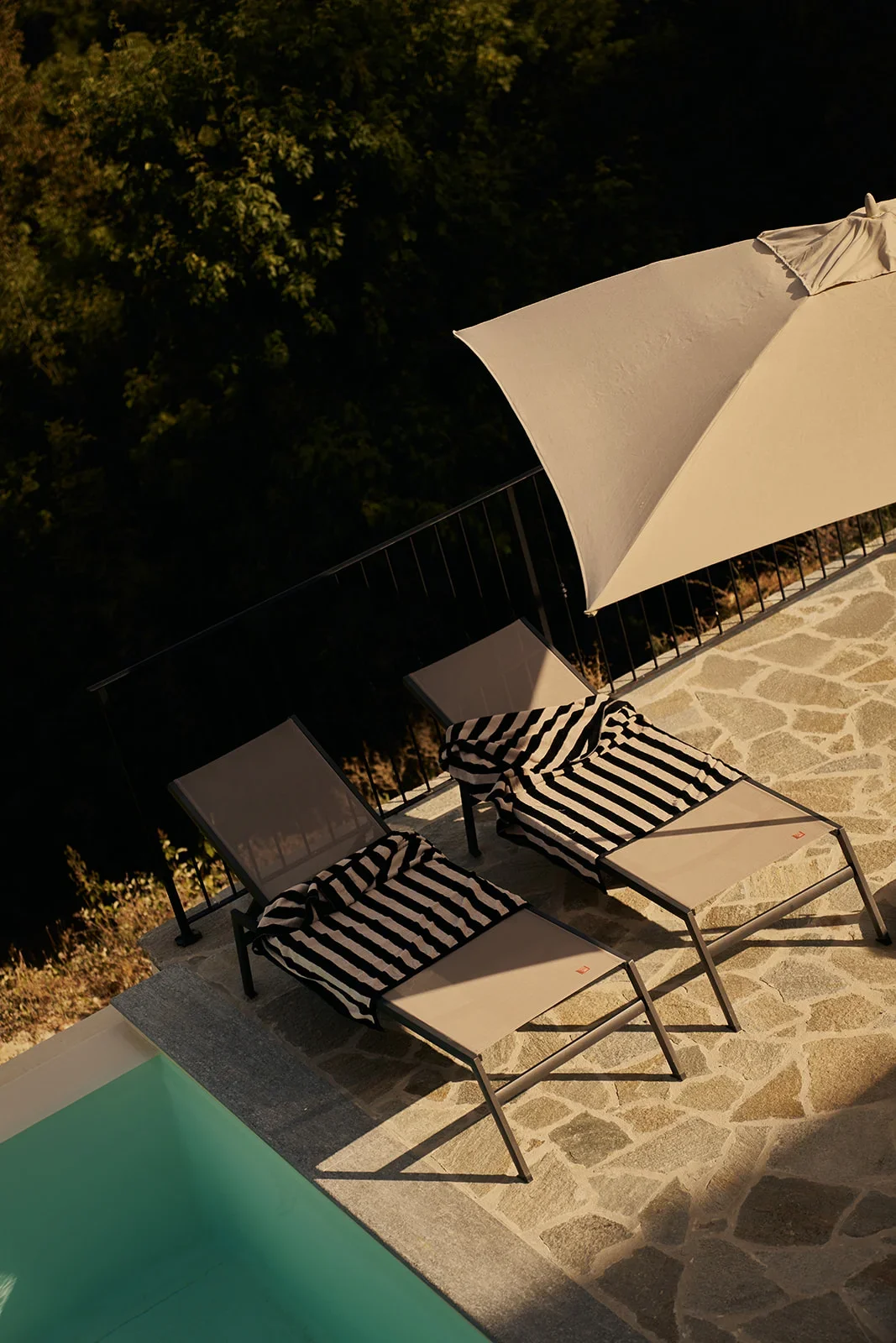 Two lounge chairs with black and white striped towels under a large white umbrella on a stone-tiled poolside patio at night.