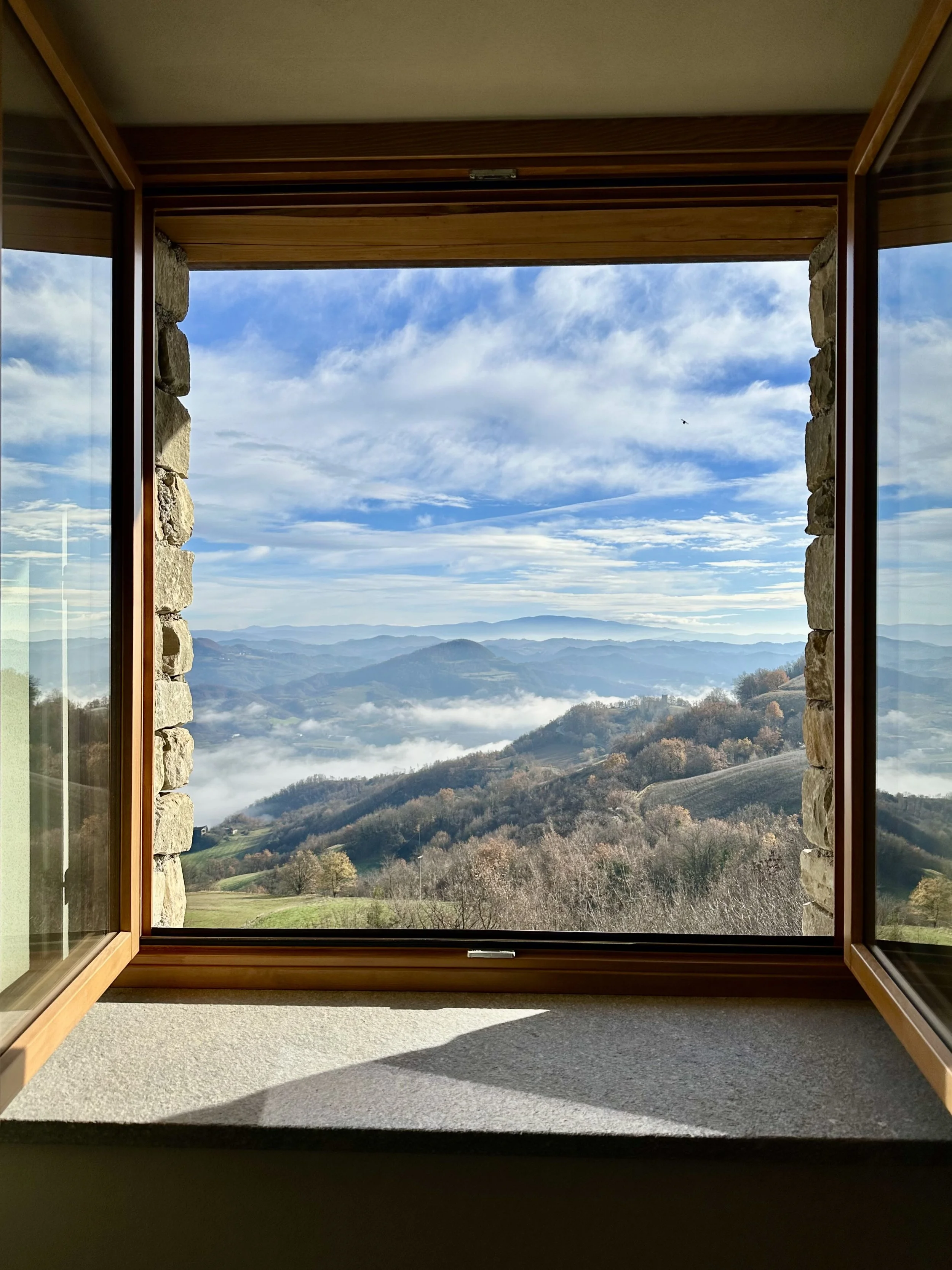 Open window with stone framing, overlooking a scenic mountain landscape with rolling hills, clouds, and distant mountains under a partly cloudy sky.