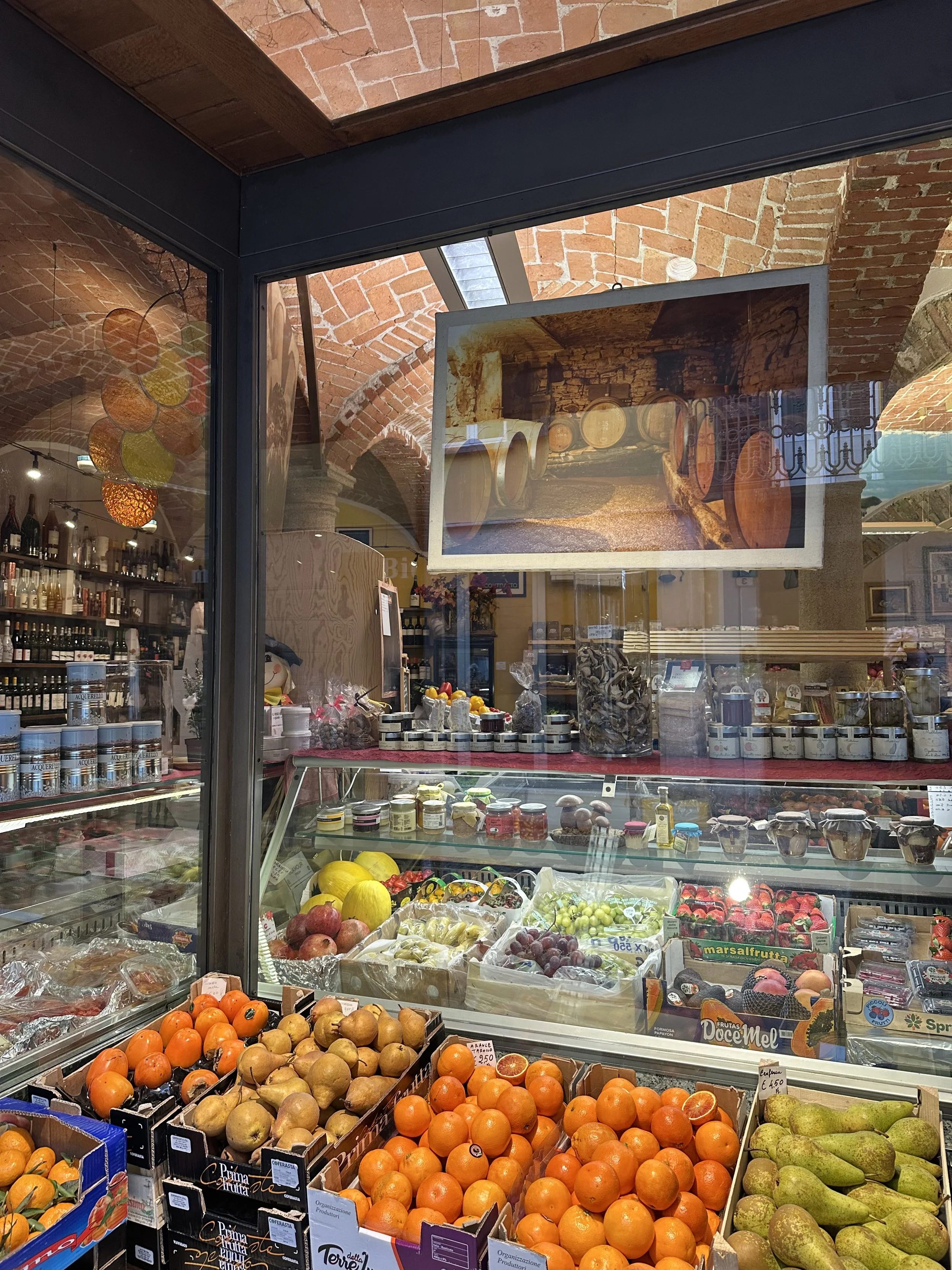 View of a fruit display inside a store with fresh oranges, pears, apples, and other produce, seen through a glass window with a reflection of a barrel room hanging on the other side.