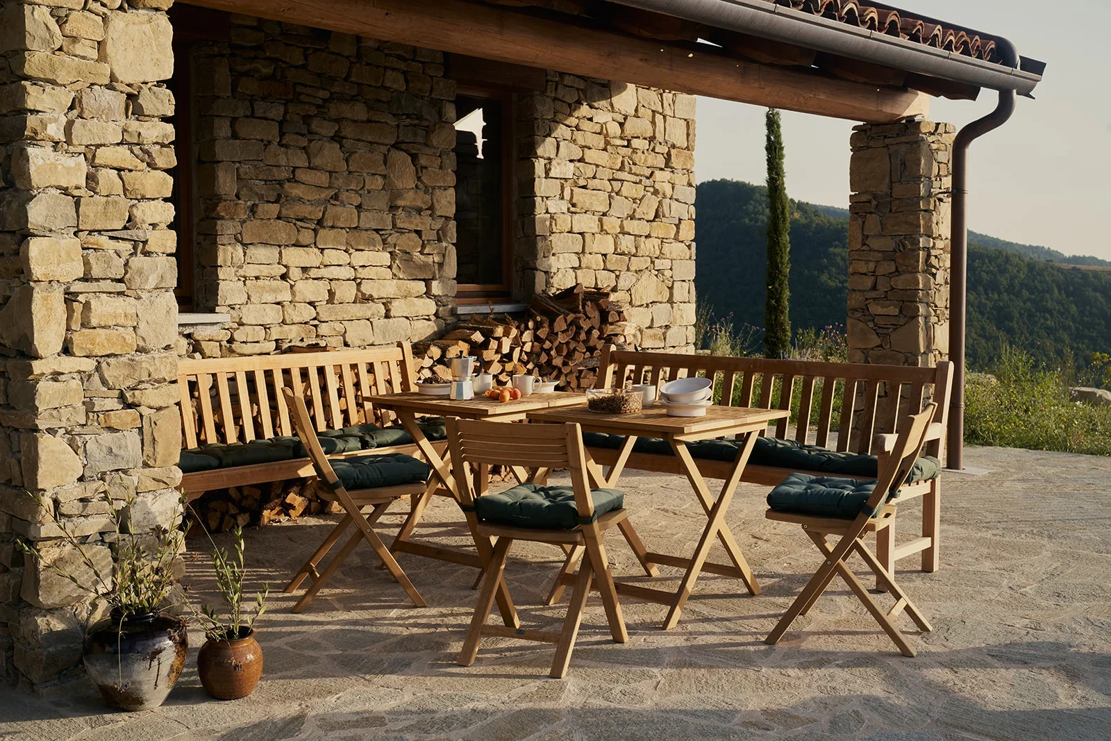 Outdoor patio with wooden table and chairs, stone house wall with stacked firewood, potted plants, scenic hilly landscape in the background.