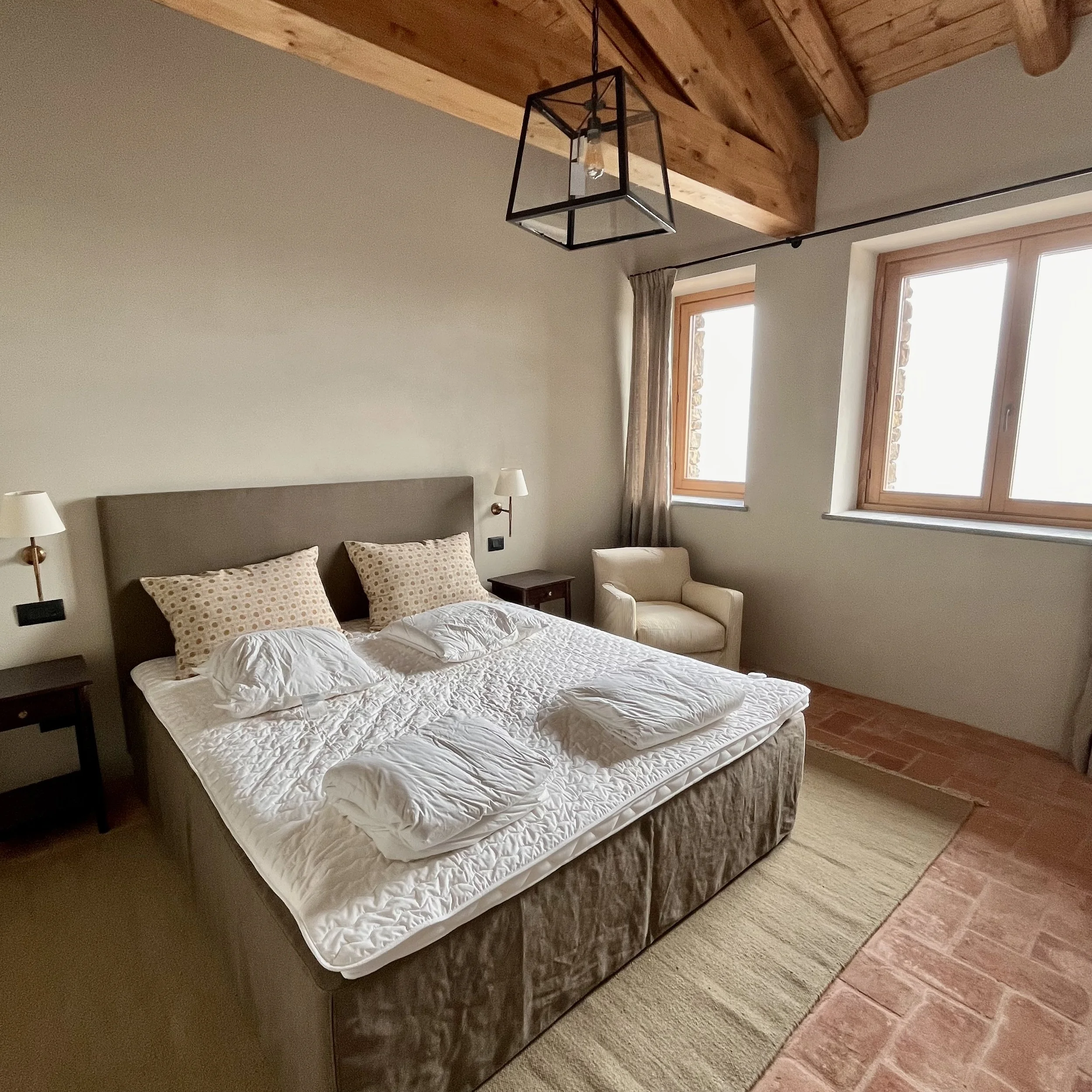 A bedroom with a large bed, two pillows, and four folded towels on top. The bed has a beige headboard and is flanked by two dark nightstands with wall-mounted lamps. There is a beige armchair next to a window, a window curtain, and two large windows 