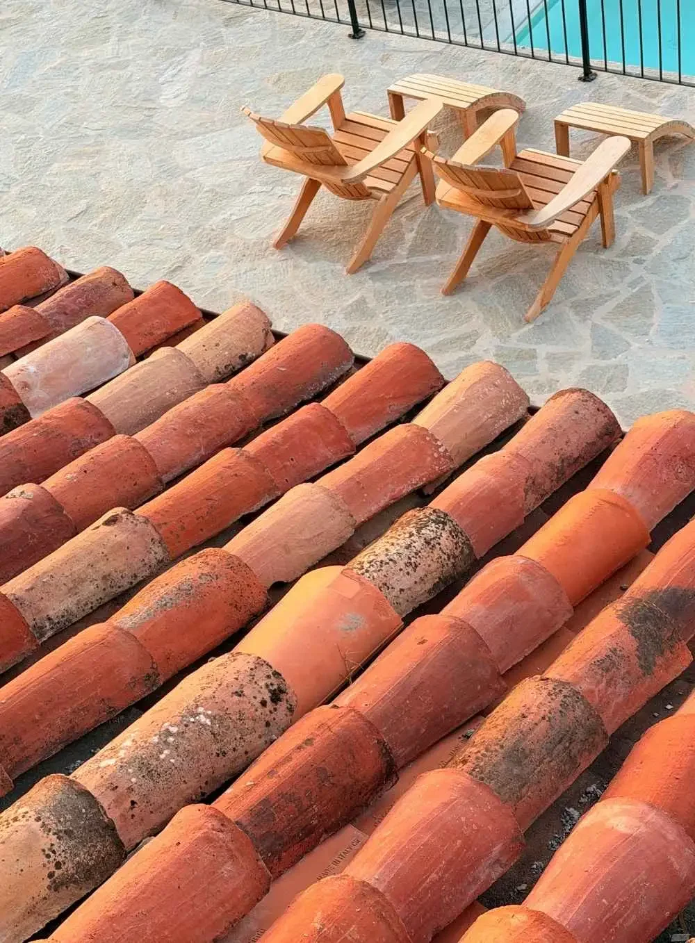Two wooden lounge chairs and a small matching table on a tiled patio next to a swimming pool, with a tiled roof in the foreground.