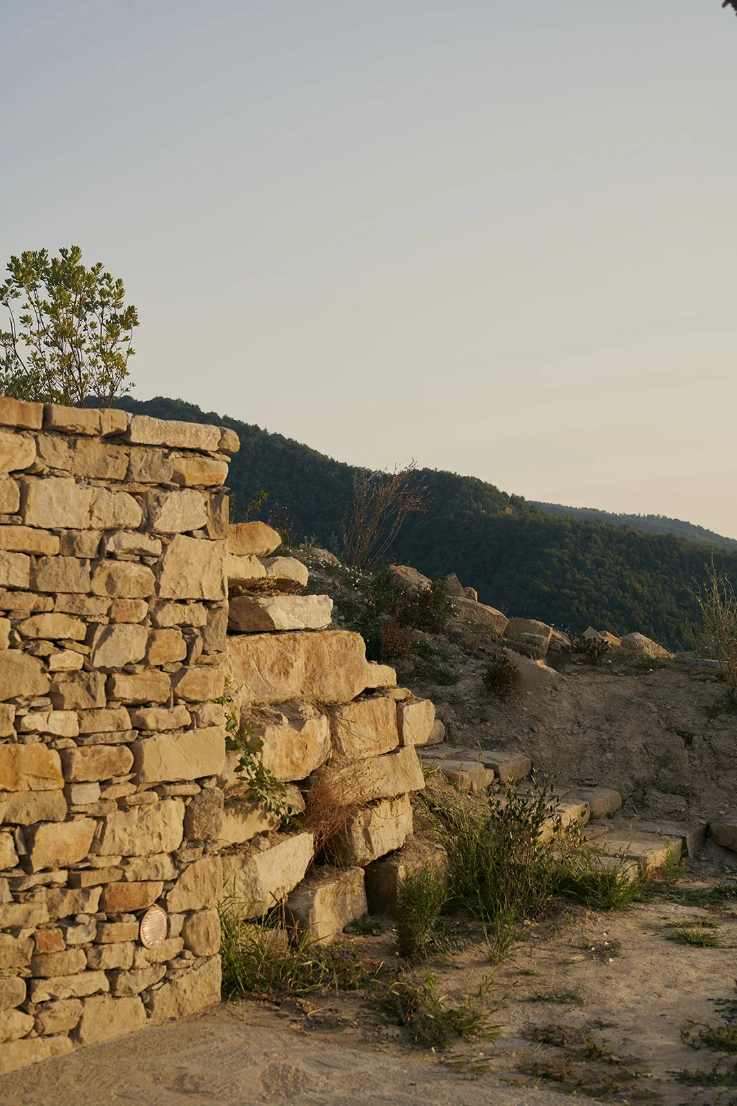 Sunlit stone steps next to a stone wall on a hillside with trees and mountains in the background.