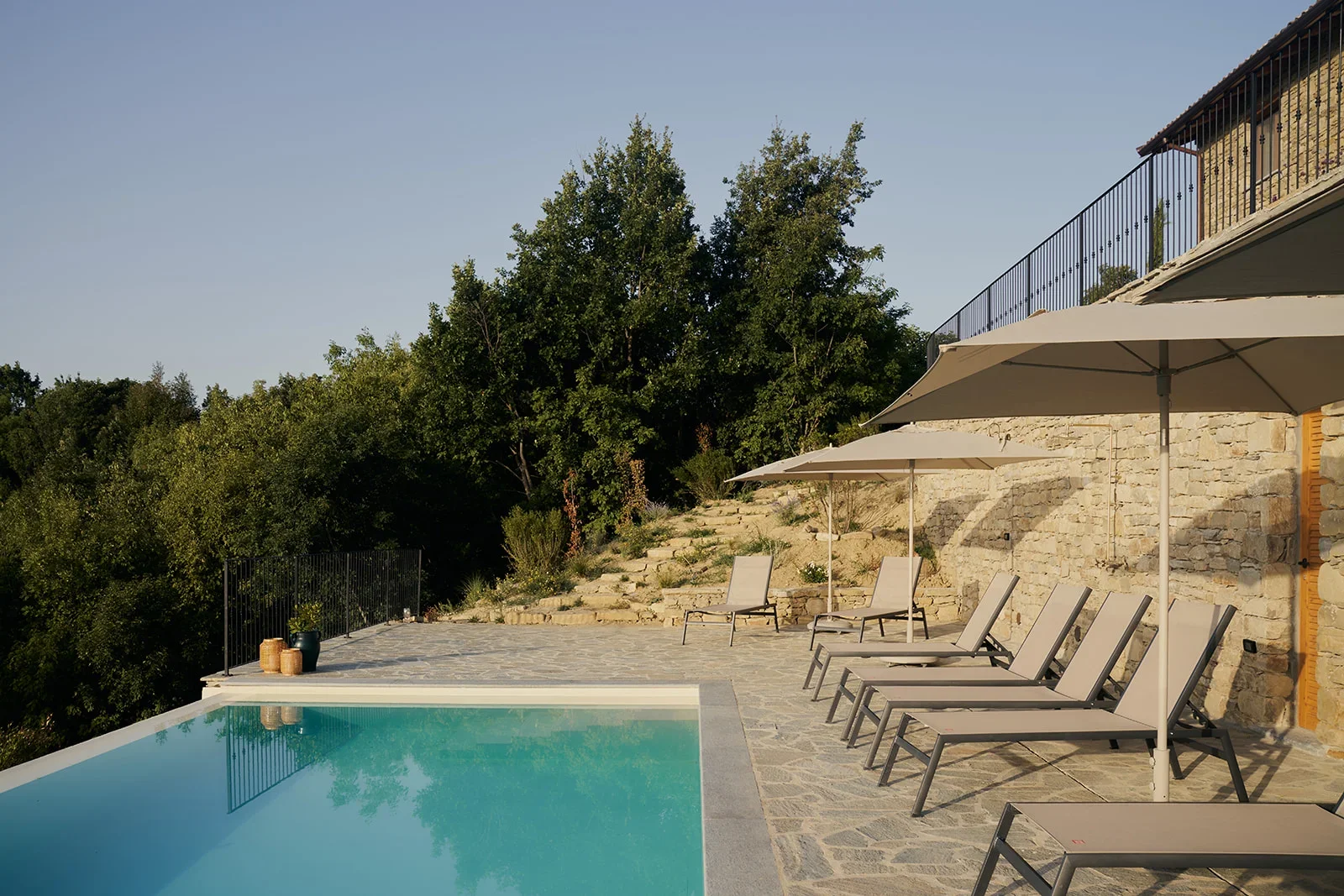 Luxury outdoor pool area with lounge chairs and umbrellas, stone patio, and lush green trees in the background during daytime.