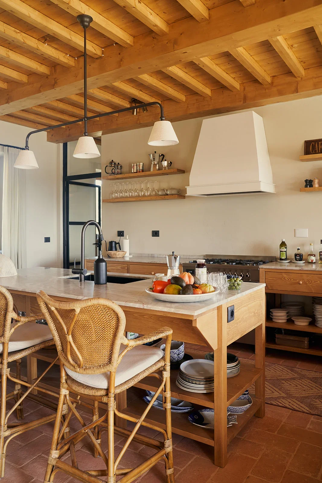 A cozy kitchen with a wooden ceiling and a large white range hood. There is a marble-topped island with a black sink and a black soap dispenser. A bowl of assorted fruits is on the island. Rattan chairs with cream cushions surround the island. Open w