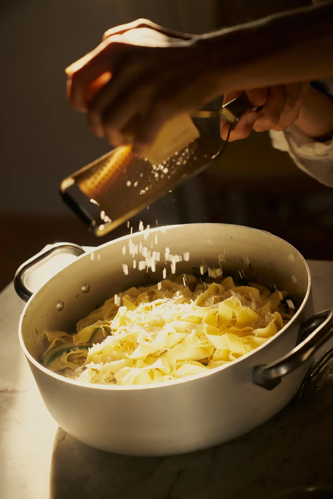 Person grating cheese over a pot of cooked pasta in a kitchen.