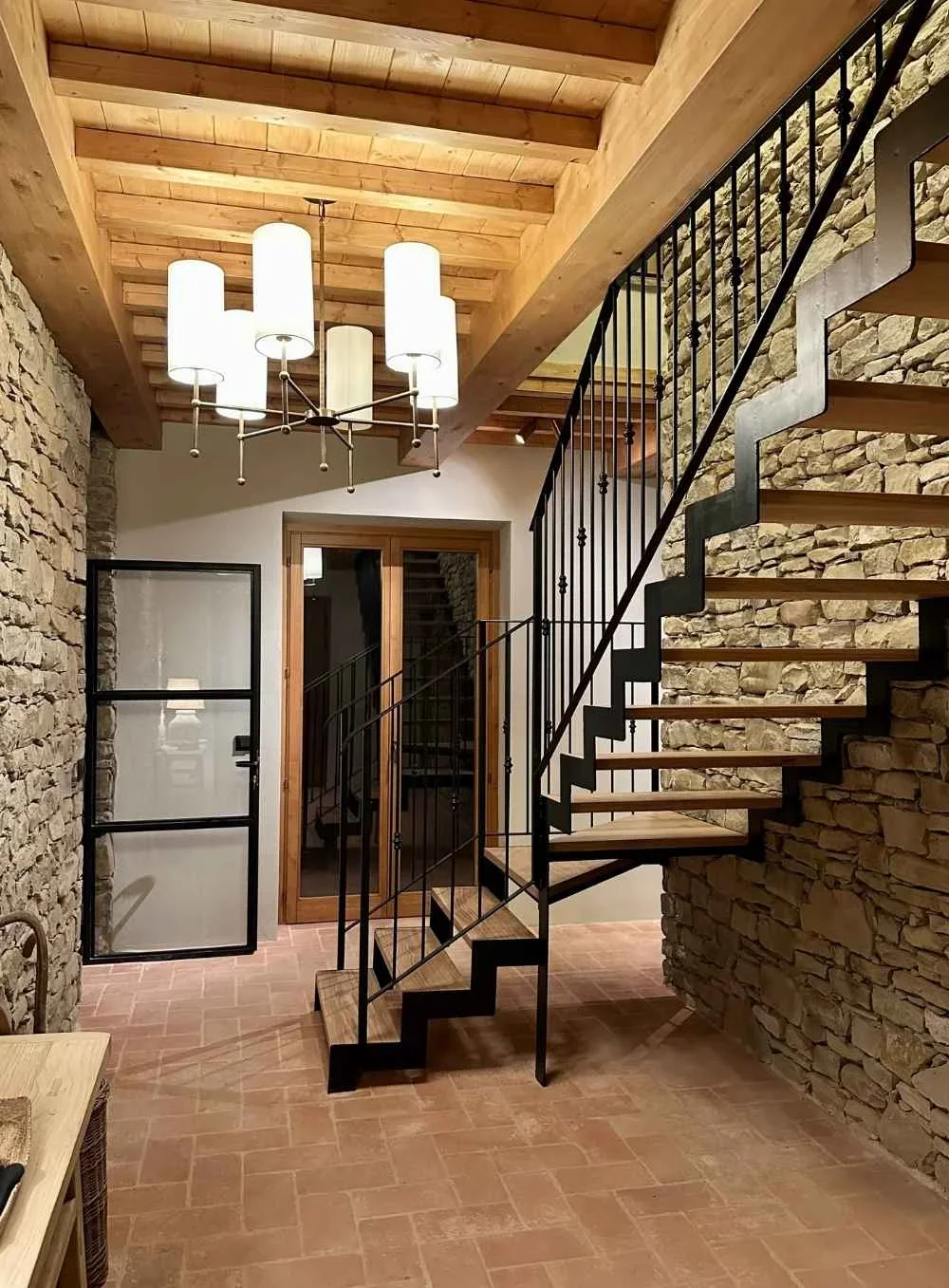 Interior of a home with a stone wall, wooden ceiling, modern chandelier, black metal staircase, and a glass door leading outside.