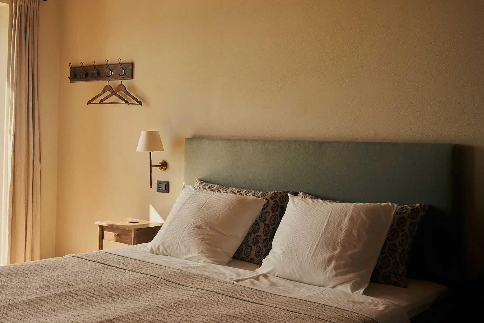 A neatly made bed with pillows in a cozy hotel room. There is a nightstand with a lamp and a wall-mounted coat rack with two hangers.