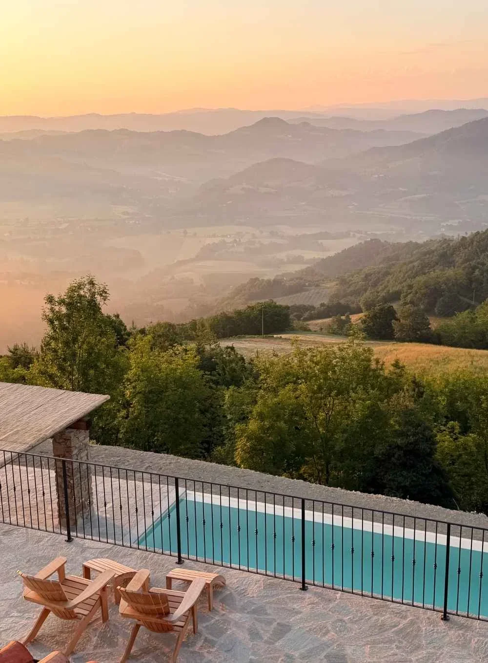 A scenic view overlooking rolling hills and valleys at sunset with a private pool and two wooden lounge chairs on a stone patio in the foreground.