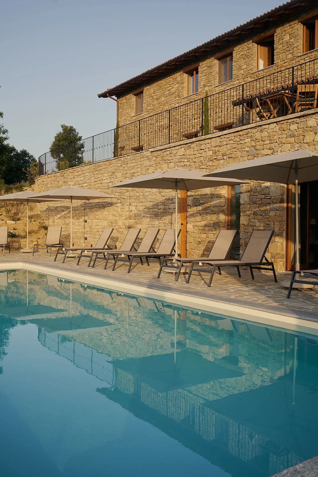 Stone house with balcony, outdoor swimming pool area with lounge chairs and umbrellas.