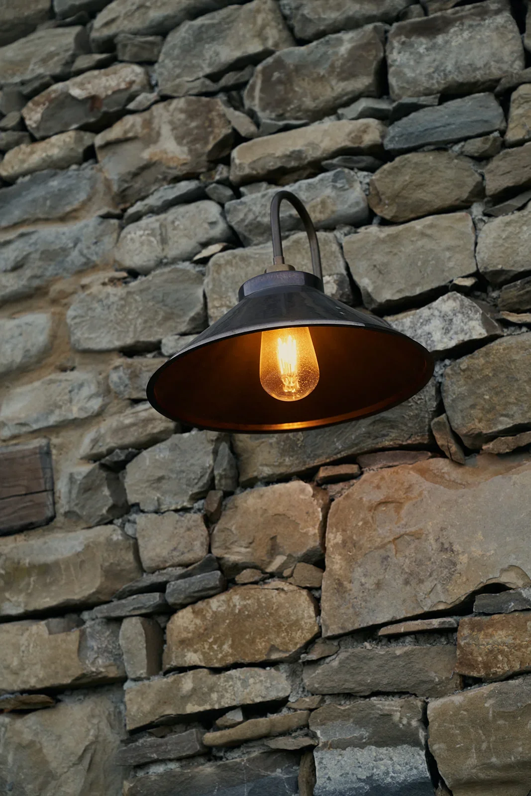 A vintage-style wall-mounted lamp with an exposed filament bulb, attached to a stone wall with uneven, natural stones.