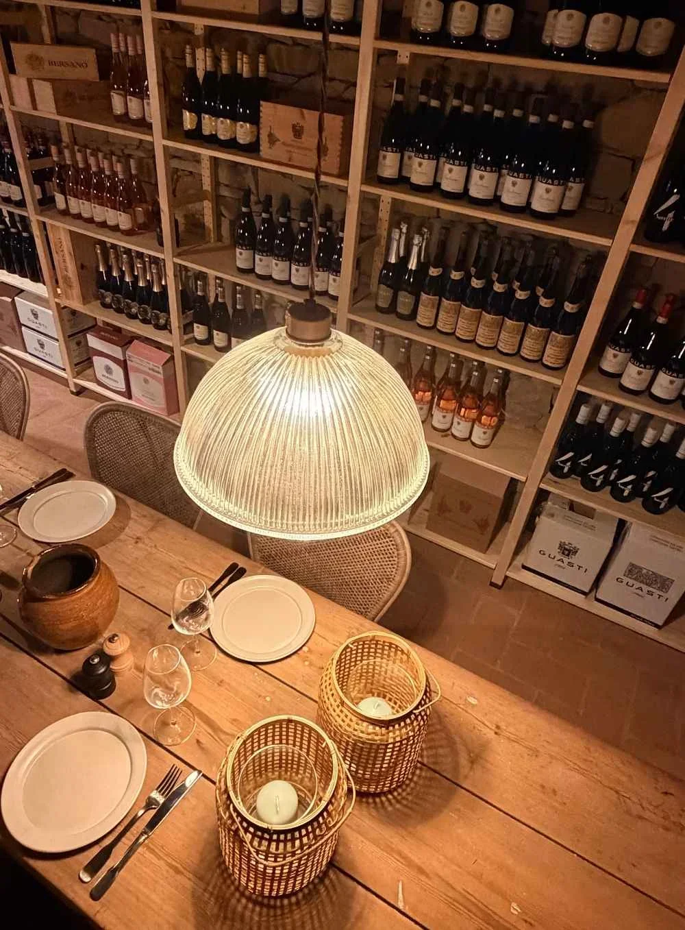 A cozy wine cellar with wooden shelves filled with bottles of wine, a rustic wooden dining table set with plates, glasses, and cutlery, and a bright pendant light hanging above the table.