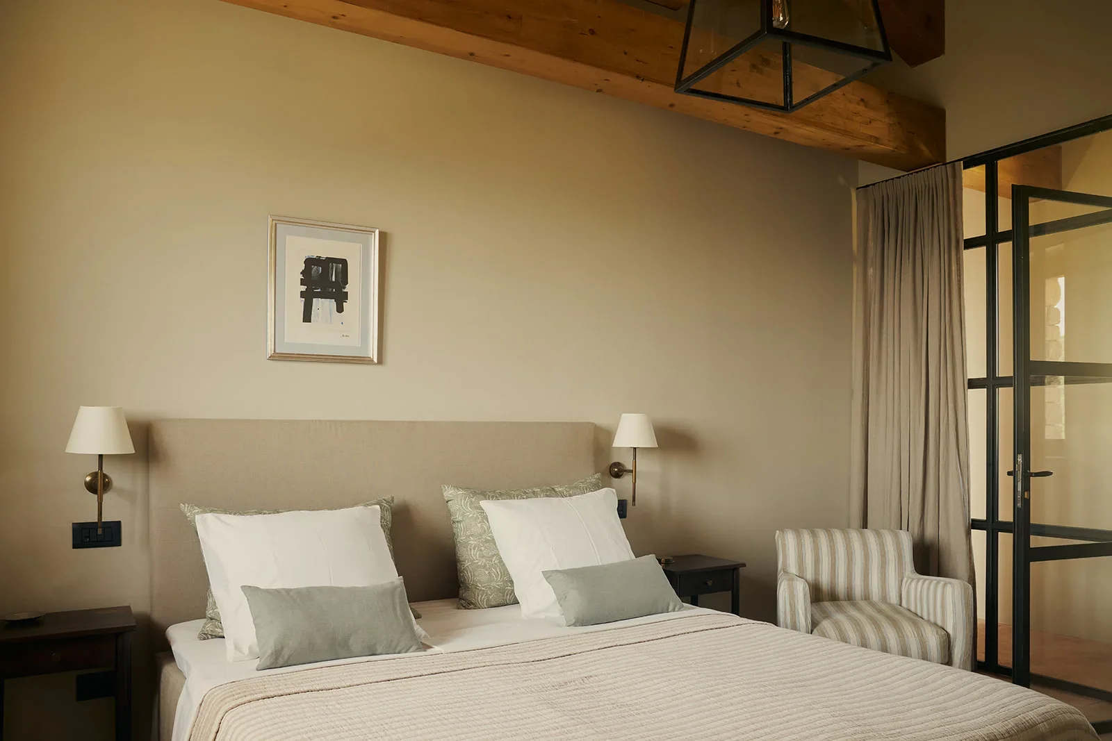 A cozy bedroom with a large bed featuring white and gray pillows and a beige headboard. On each side of the bed, there are small black nightstands with lamps. The wall above the bed has a framed black and white art piece. There is a striped armchair 