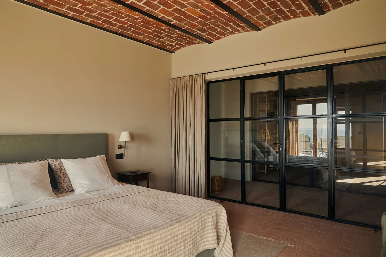 A hotel room with a bed, a nightstand, and a wall-mounted lamp. The room has a brick ceiling and a large glass sliding door with black frames, leading to an outdoor balcony.