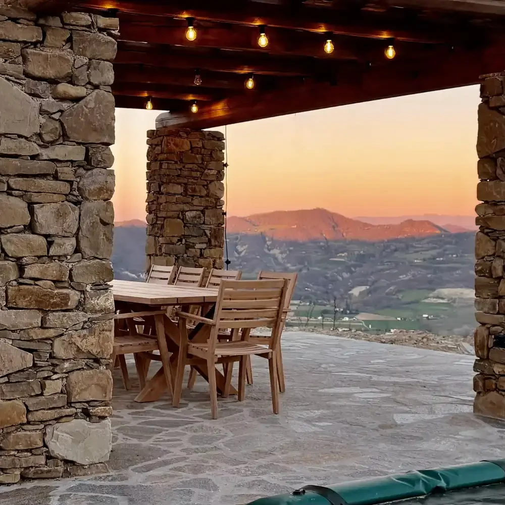 An outdoor stone patio with a wooden dining table and chairs, overlooking a mountainous landscape at sunset, with string lights hanging from the wooden ceiling.