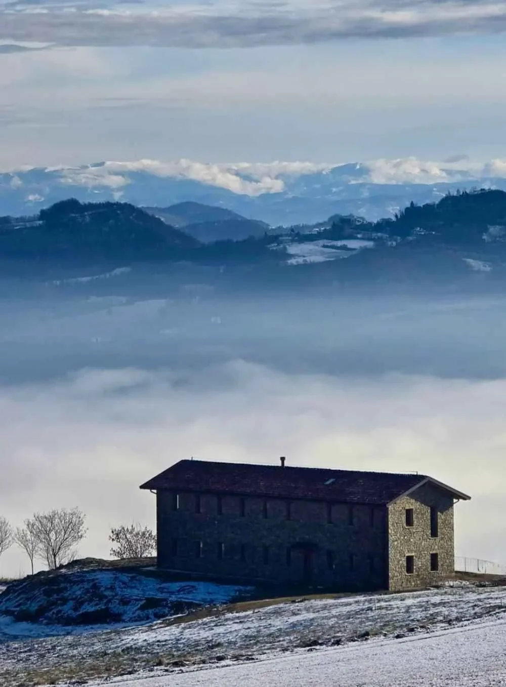 A stone house on a snowy hilltop surrounded by fog and overlooking a mountain range in the distance.