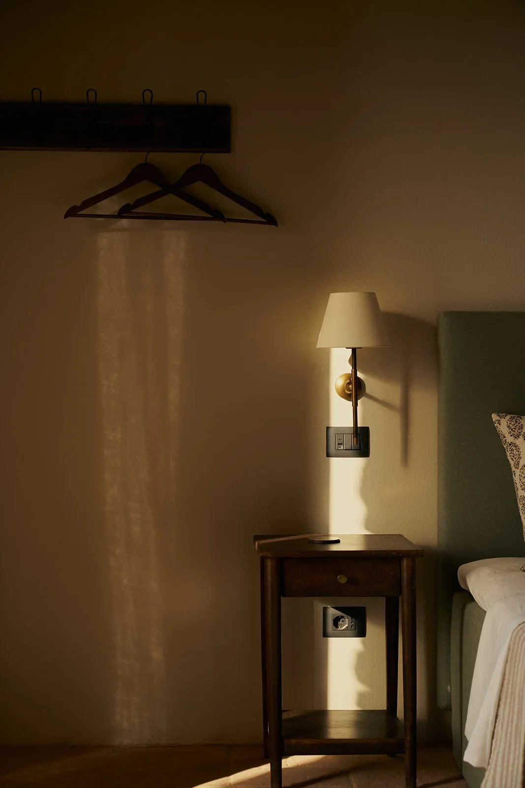 A bedside table with a remote control, a wall-mounted lamp casting shadows, an outlet, and part of a bed with a patterned pillow in a dimly lit room.