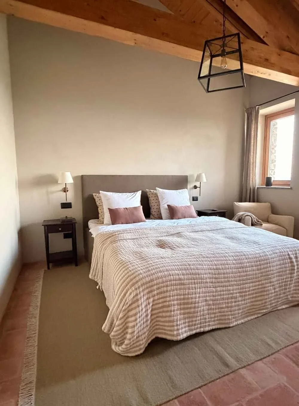 A cozy bedroom with a large bed, beige striped bedding, and decorative pillows. There are two matching bedside tables with wall-mounted lamps, an armchair with a throw blanket near the window, and a modern geometric pendant light hanging from the woo